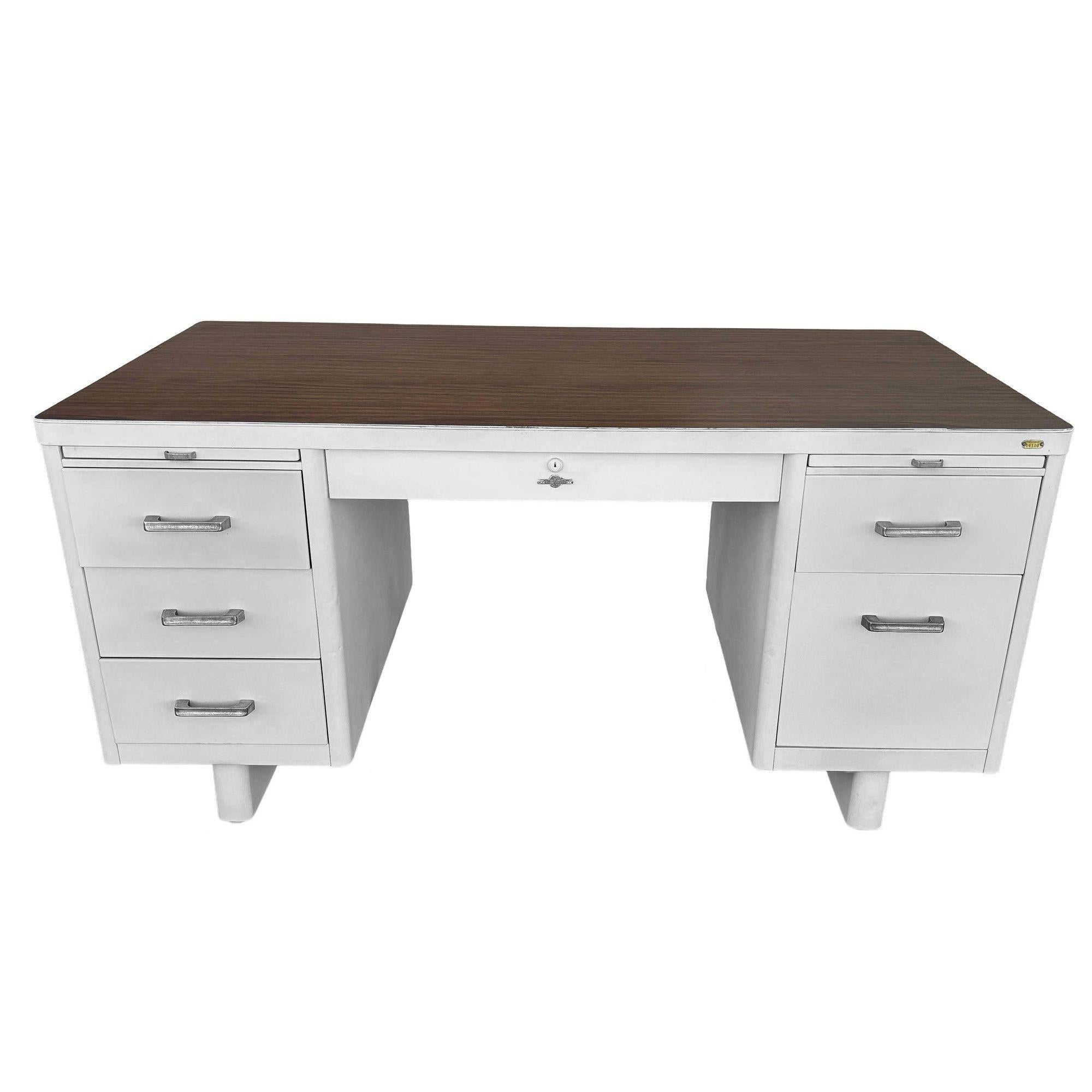 La scrivania per cisterna in acciaio verniciato bianco Atomic Age presenta un piano in Formica con venature del legno, 2 tavoli da lavoro estraibili e un cassetto centrale con 5 cassetti laterali. La scrivania è realizzata in acciaio di grosso