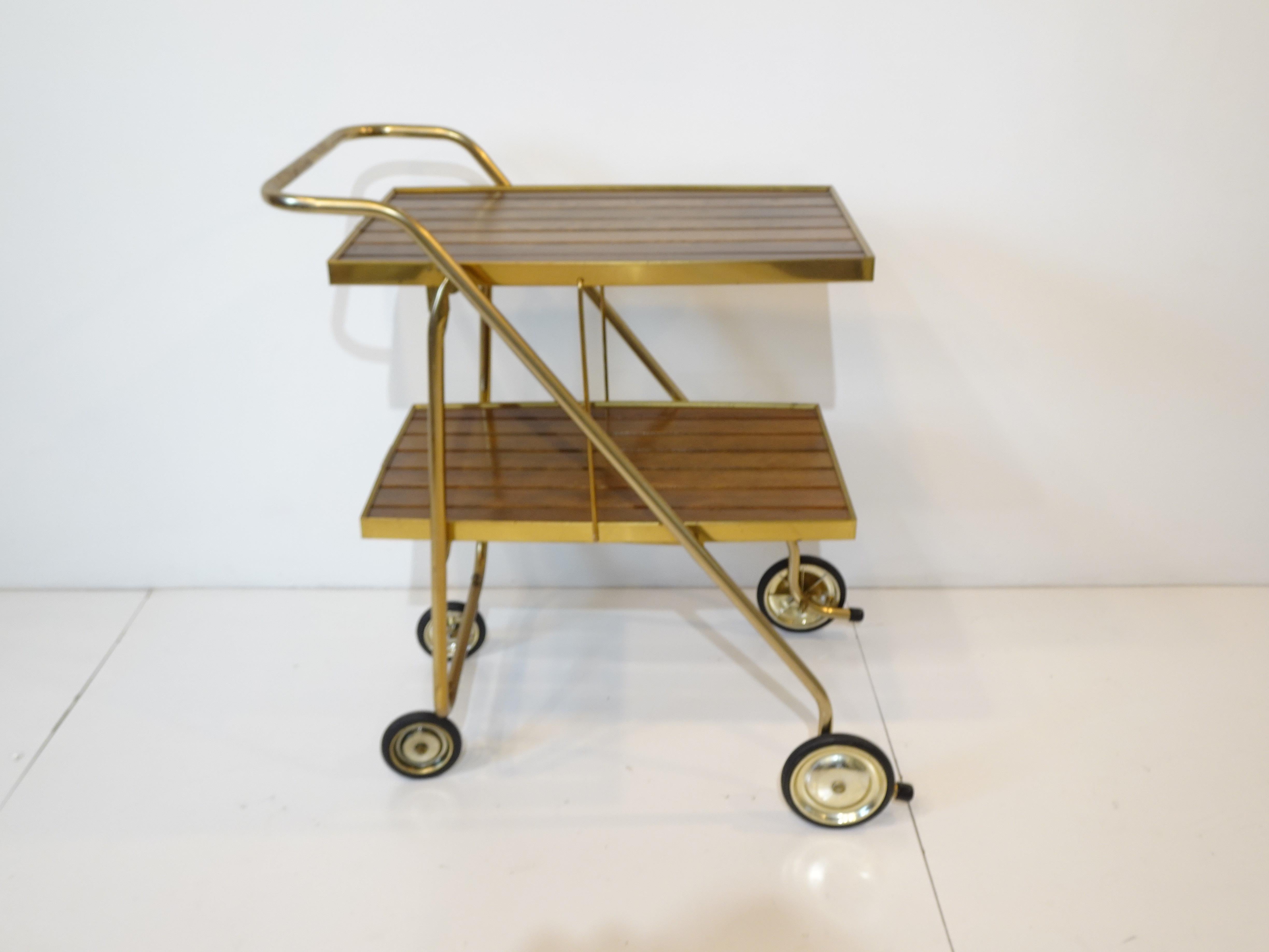 A brass toned metal framed folding bar cart trolley with darker wood slat top and bottom shelve having wheels with the rear ones that swivel for easy movement. This well thought out design folds tightly and compactly for storage or set up is very