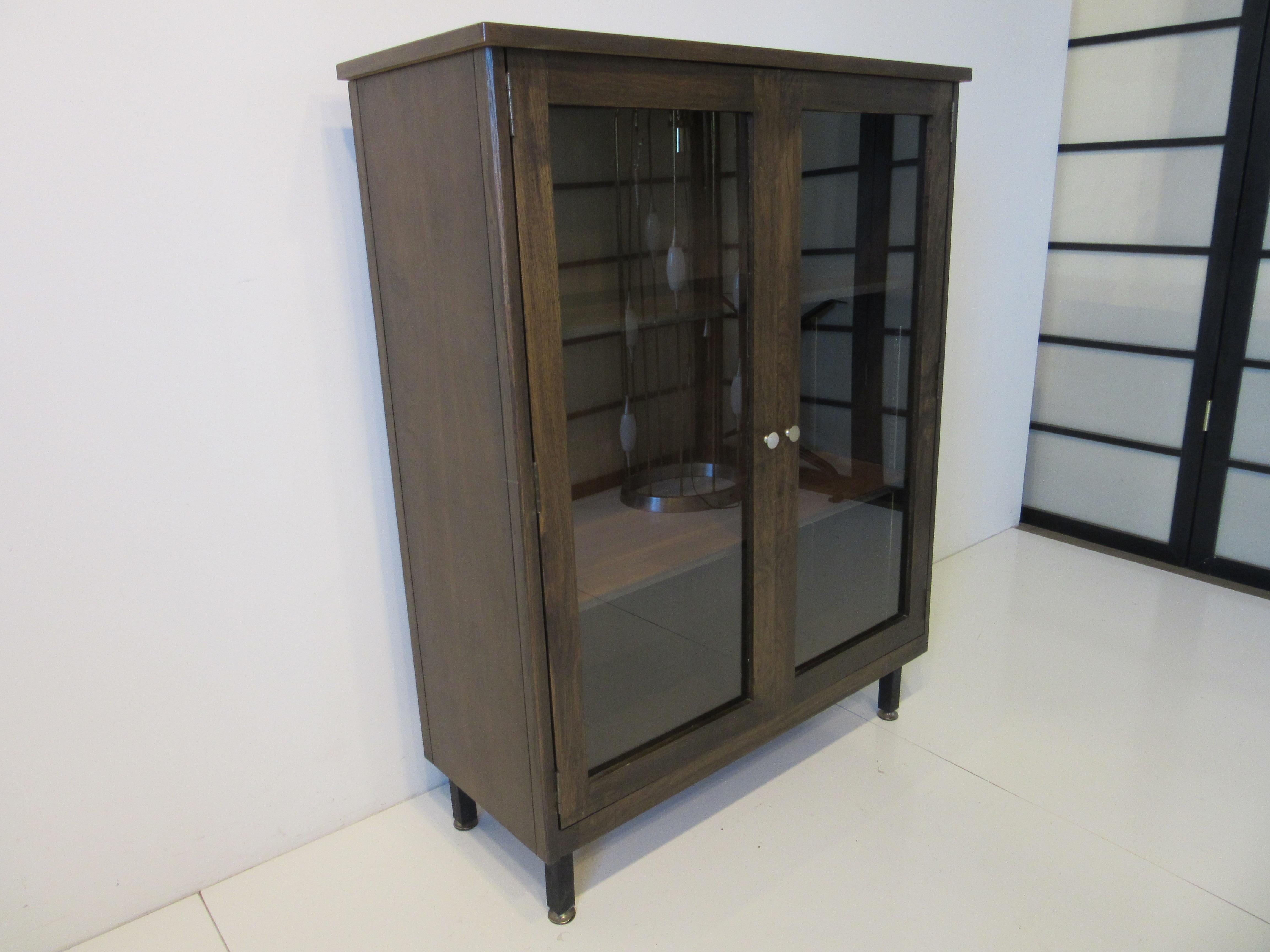 A grained wooden bookcase with one fixed shelve and one adjustable shelve sitting on very sturdy satin black squared tubed legs with adjustable foot pads . The pulls on the double doors are in brushed aluminum so it has that Mid Century vibe but
