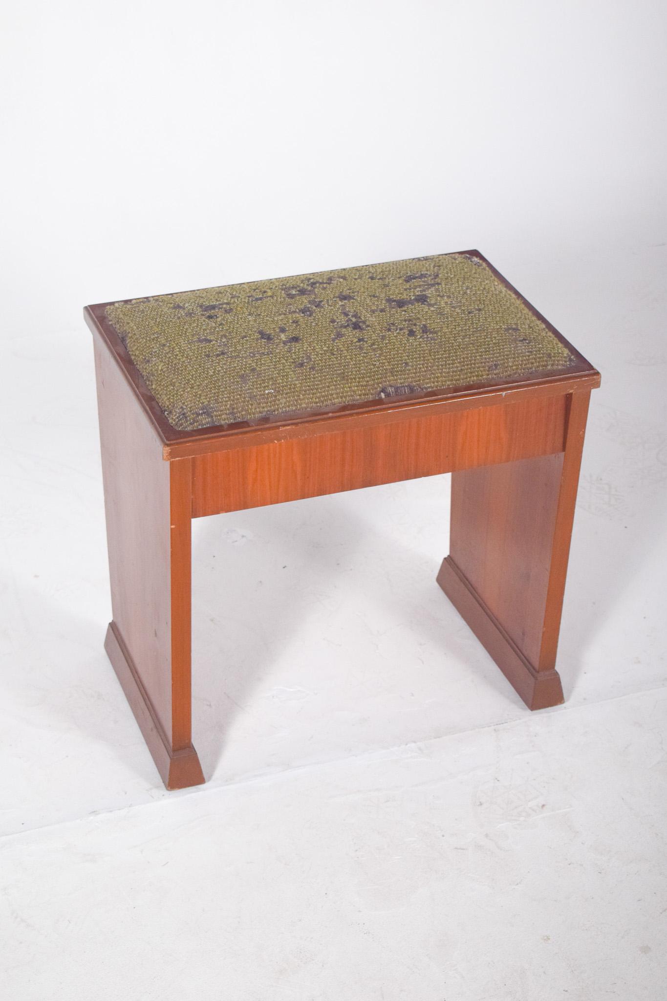 Banc de piano en bois. Avec une finition en vernis clair, des lignes droites et une structure robuste, caractéristiques des meubles fonctionnels du milieu du XXe siècle. Le siège rembourré, actuellement recouvert d'un tissu vert usé, présente des