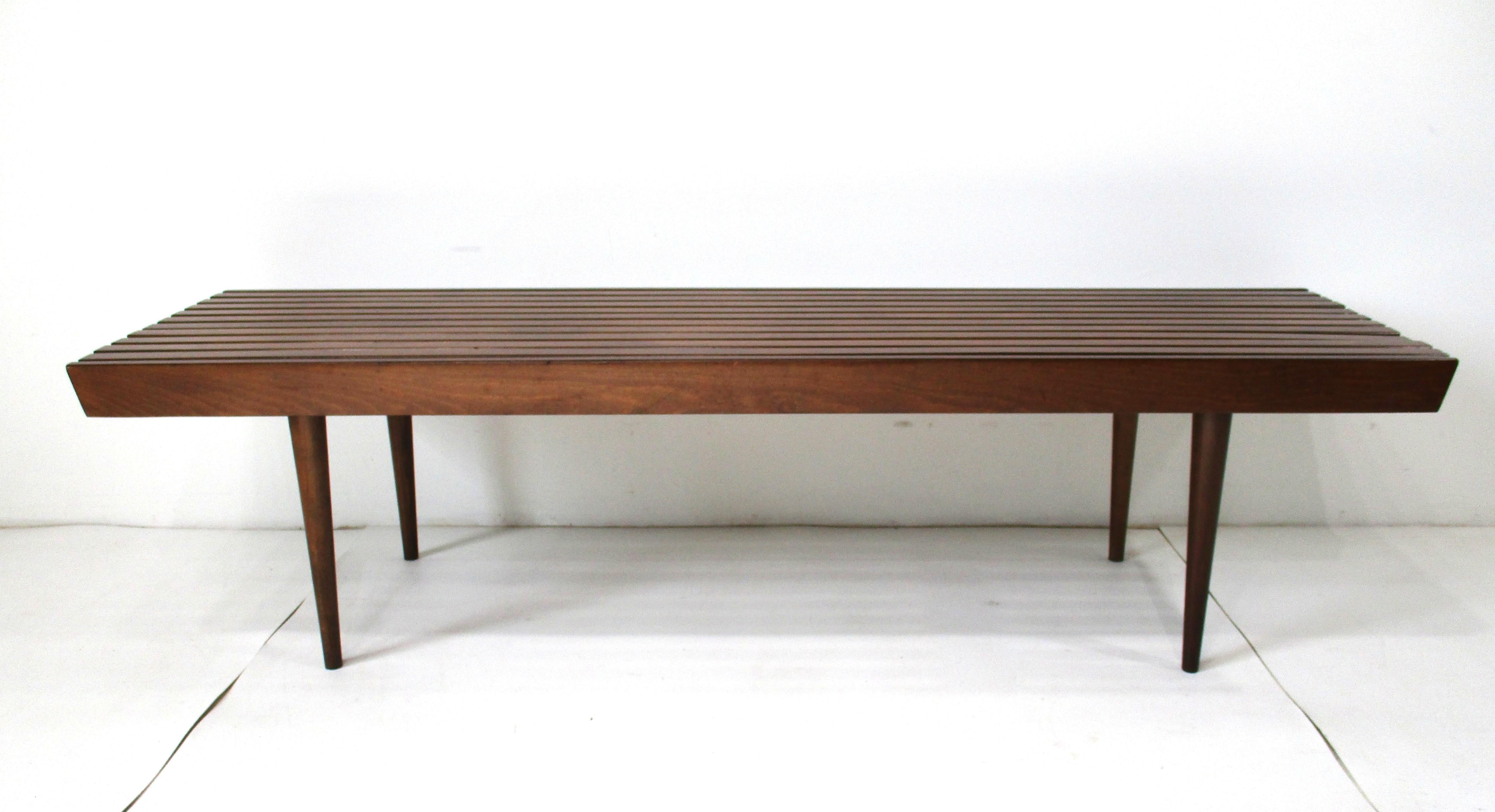 A Mid Century walnut toned slat bench or coffee table with conical legs and tapered ends. This simple design is still as fresh today as it was back in 1950 when furniture was versatile being crafted for different uses as the family expanded, bought