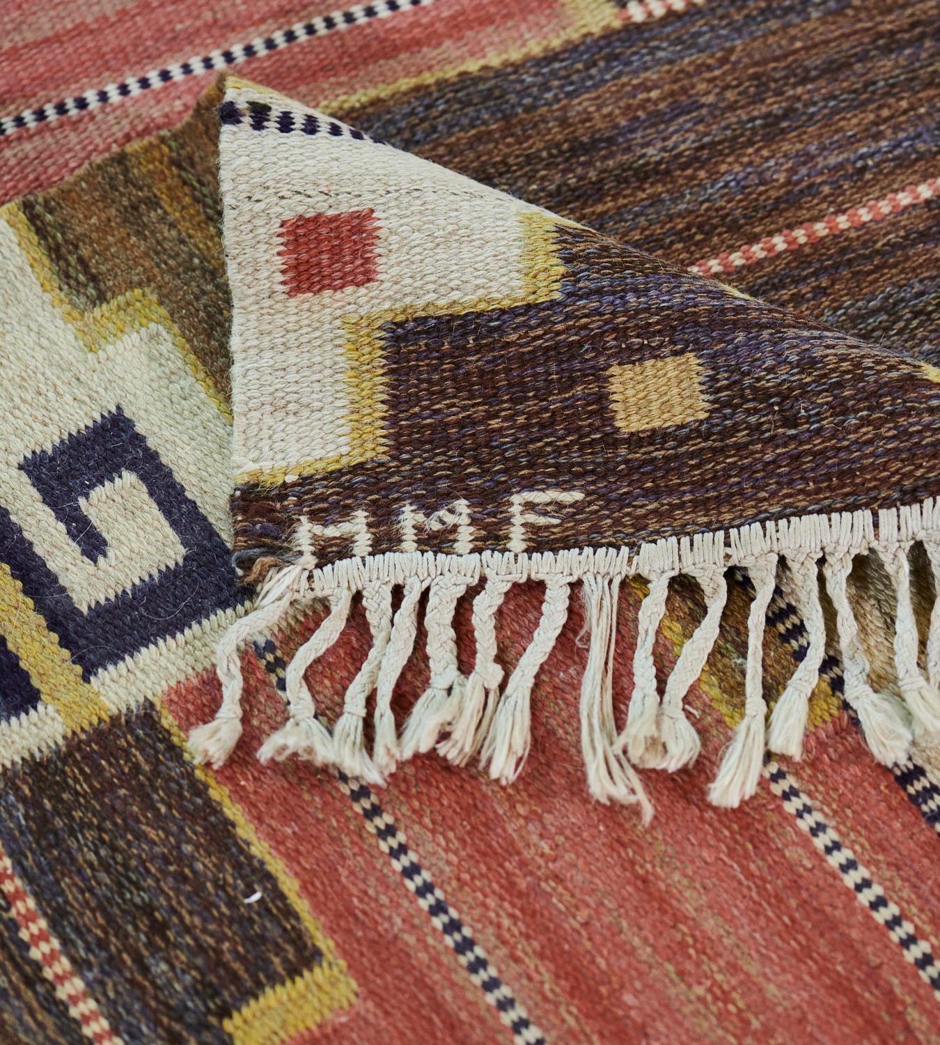 This mid-century signed Swedish rug has a field with shaded terracotta, chocolate-brown, sandy-brown and light burgundy-red stepped and linked lozenges outlined with a yellow stripe, horizontal rows of tiny ivory and black checkerboard stripes, in a