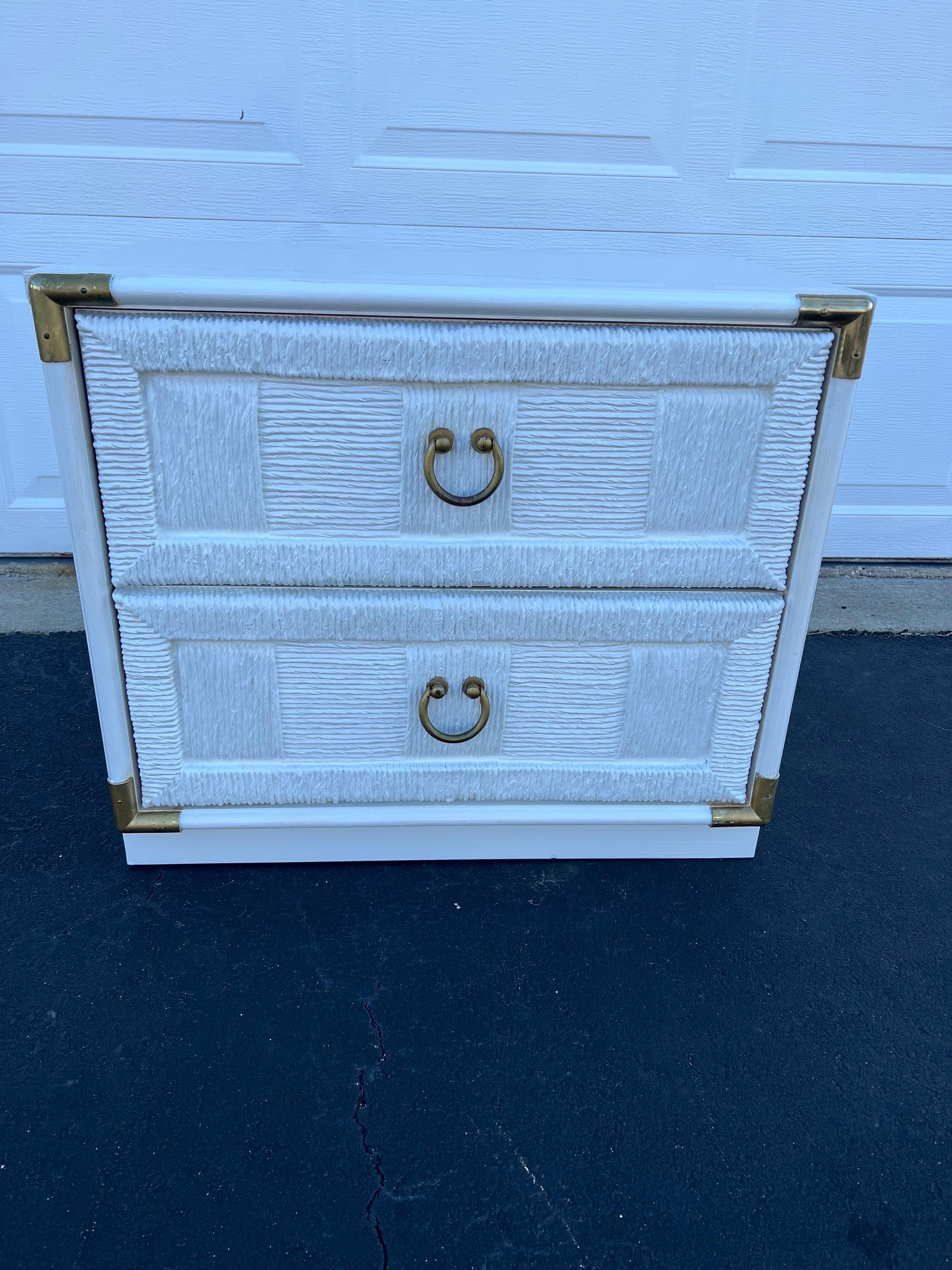 Mid Century Woven Nightstand in White. Nice coastal two drawer piece with laminate top. Chunky brass hardware frames this chest with a classic campaign feel. The textural front make this piece a stand out. Use as a nightstand or as an end table.