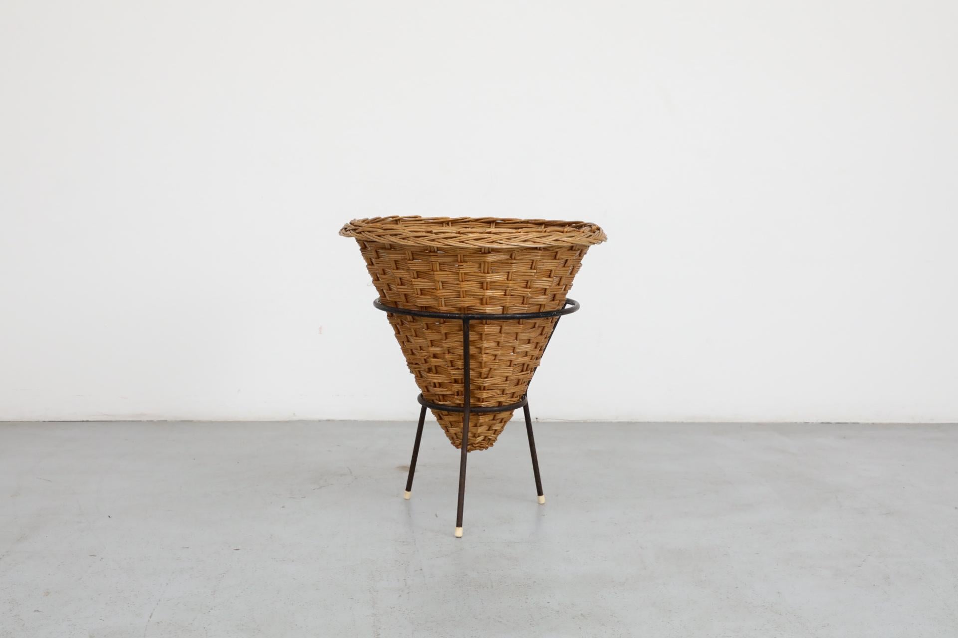 Cestino in rattan intrecciato di metà secolo con telaio a treppiede smaltato nero. Ottimo per coperte, riviste, piante o biancheria. In condizioni originali, con un'usura visibile che comprende la rottura del rattan e la perdita di smalto. L'usura è