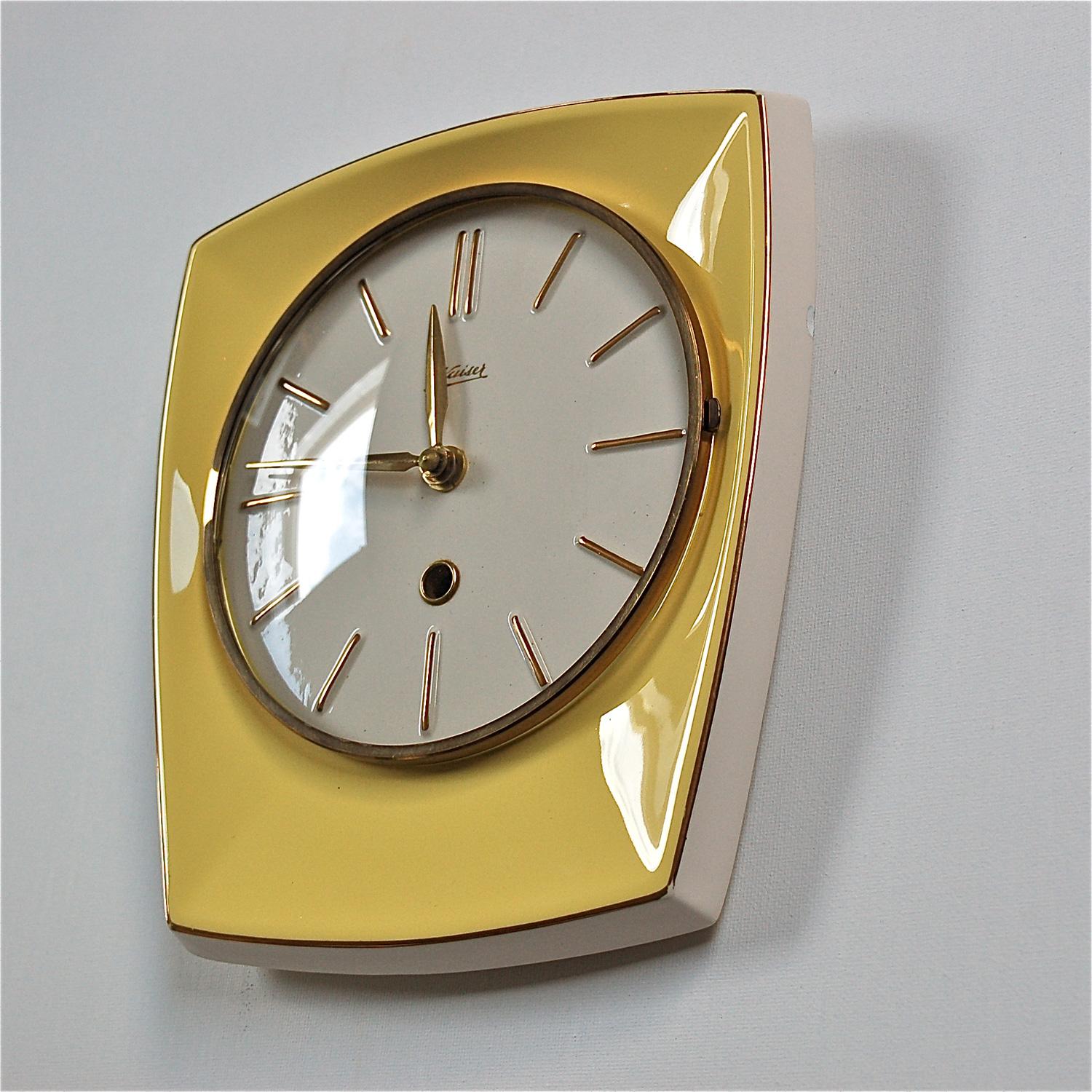 1950s vintage ceramic wall clock by Kaiser. The circular clock face sits is a rectangular base with curved edges. The ceramic base is cream colored with a custard yellow glazed front. The clock has a hinged glass front with a thin brass bezel. The
