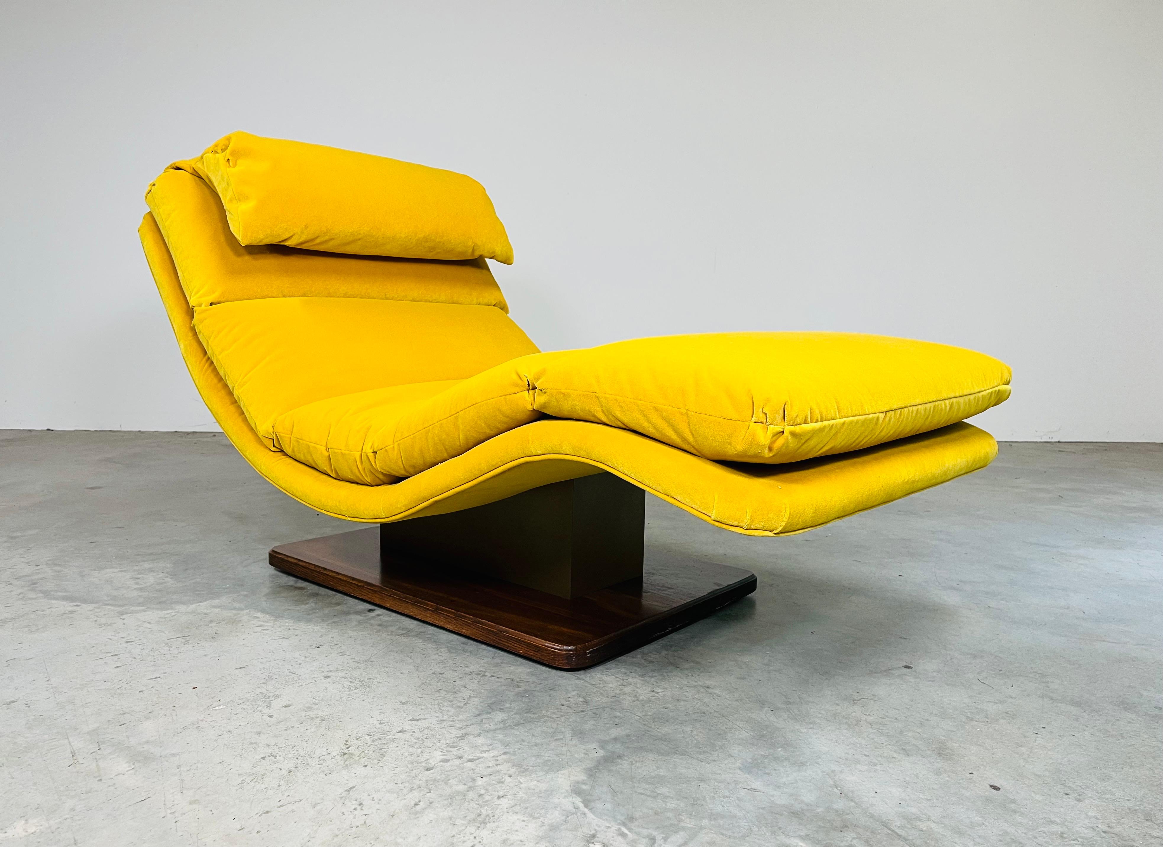 An incredibly comfortable wave chaise lounge chair by Carsons having natural wooden base with brass toned steel spacer under a golden yellow velvet lounge seat that features channeled seat cushions and a flip over headrest. In excellent condition
