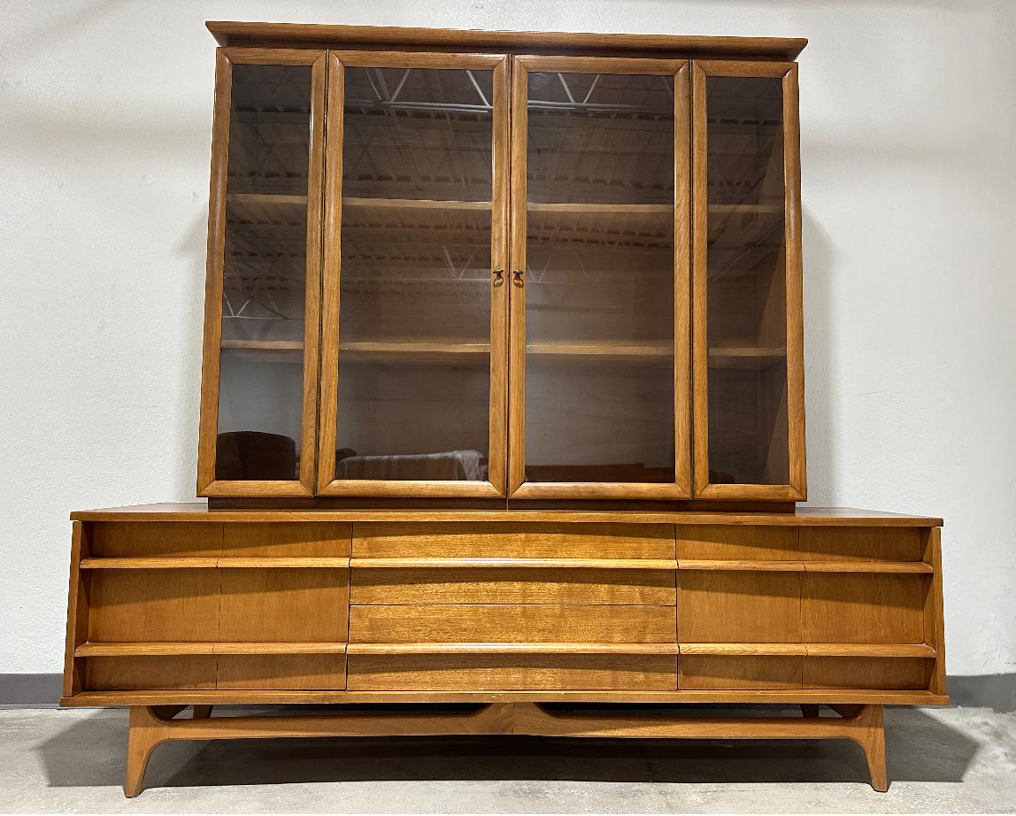 Mid Century Modern Two Piece China Hutch display cabinet by Young Manufacturing. Curved front lower credenza with carved pulls running length of front. Two drawers center, flanked by bi-fold doors with piano hinge revealing open storage. Topper has
