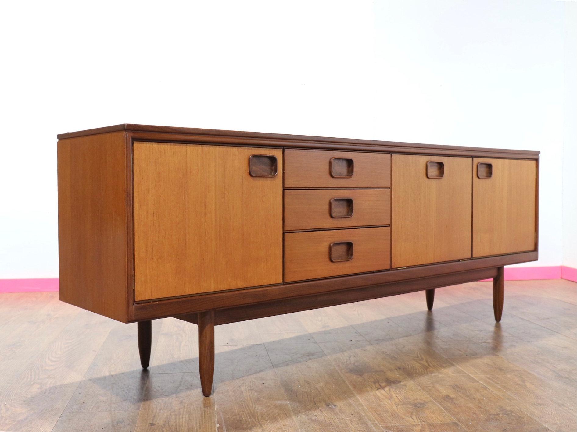 At Mid Century Traders, we proudly present a beautiful William Lawrence of Nottingham teak sideboard, with low gallery back over three short drawers and a fall front, flanked by two cupboard doors, enclosing shelves, raised on tapering cylindrical