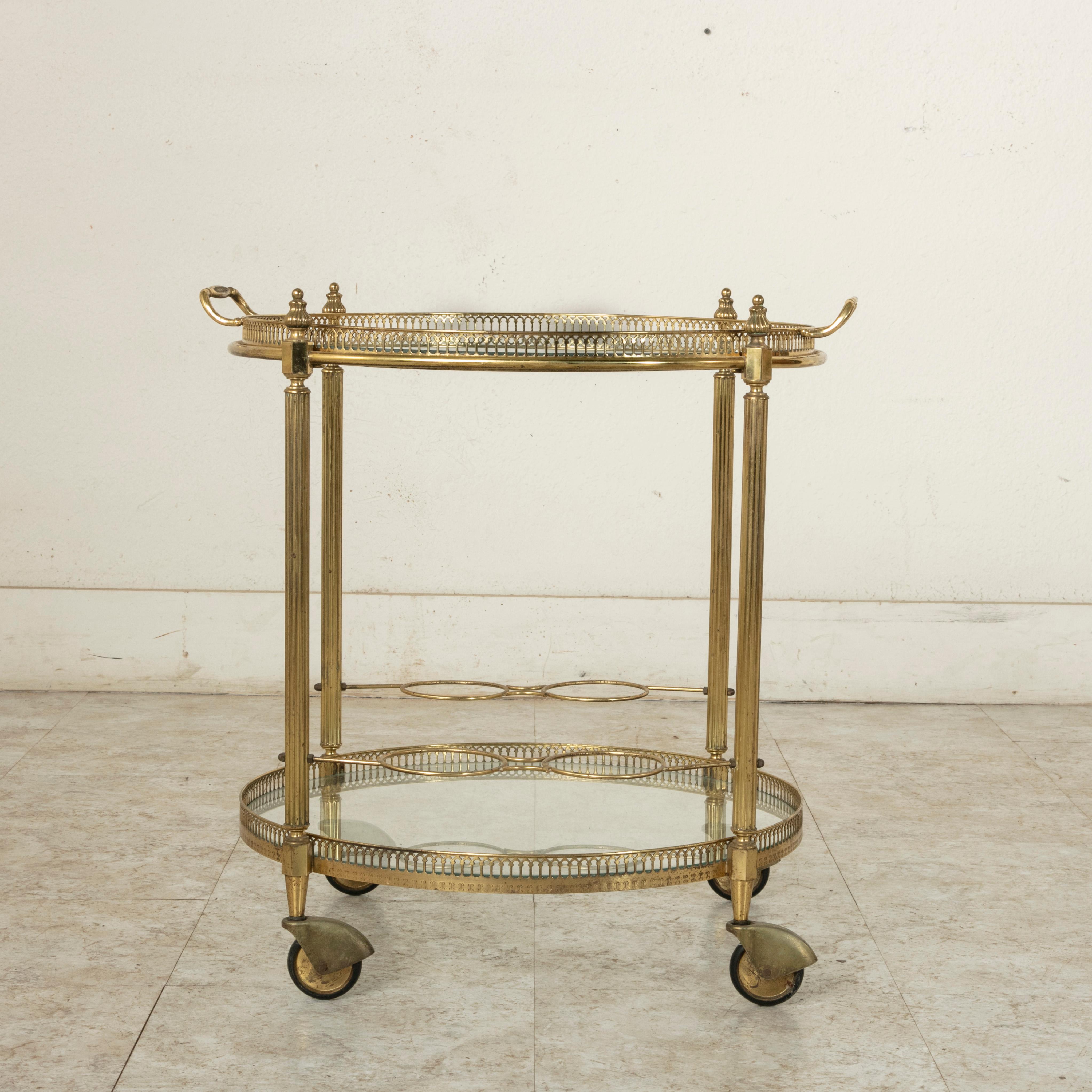 This mid-20th century French round brass bar cart features fluted legs finished with Louis XVI inspired finials and two glass shelves surrounded by a pierced bronze gallery. The upper shelf also serves as a removable serving tray with two handles to