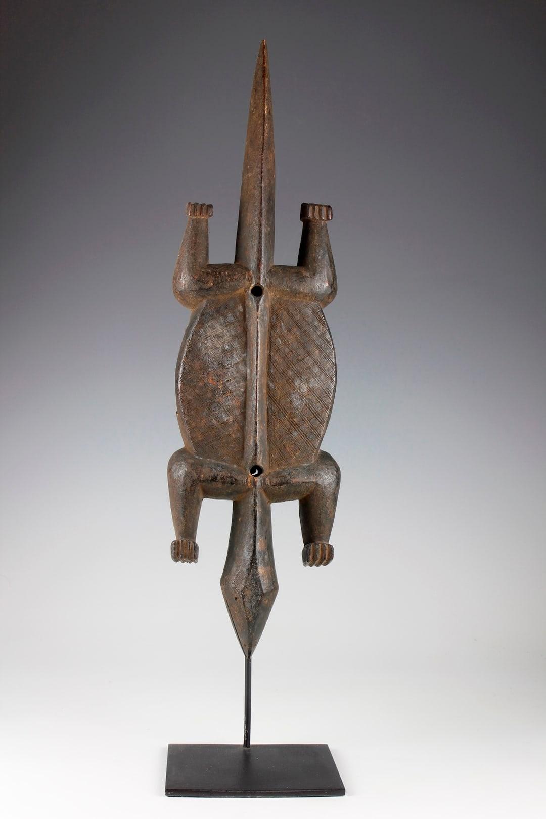 Questa serratura Bambara della metà del XX secolo, proveniente dal Mali, è stata intagliata a forma di coccodrillo, simbolo di protezione nella cultura Bambara. La serratura della porta a forma di coccodrillo, ornata da disegni incrociati finemente
