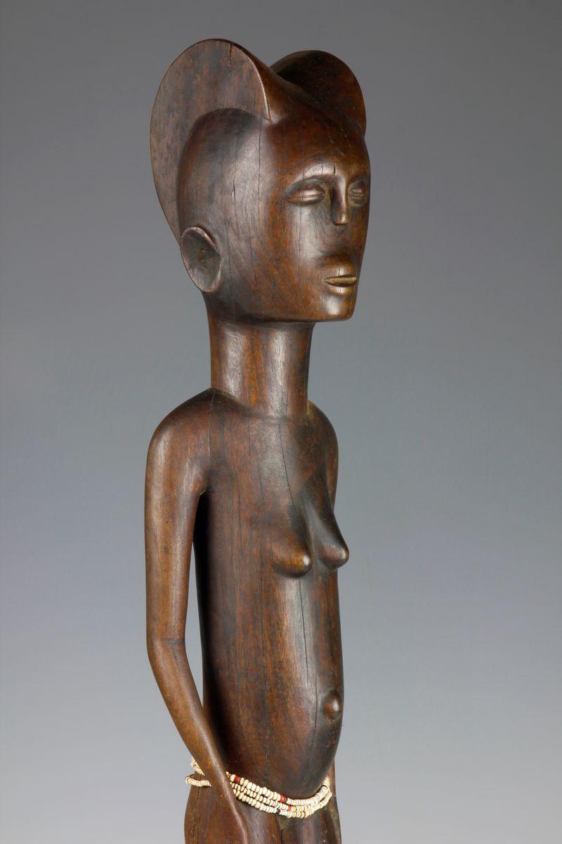 This mid-twentieth century figurative sculpture, which comes from the Kwere culture in Tanzania, depicts a female standing in an upright stance, with her hands placed by her sides. 

A prominent parted carved coiffure crowns her head, and