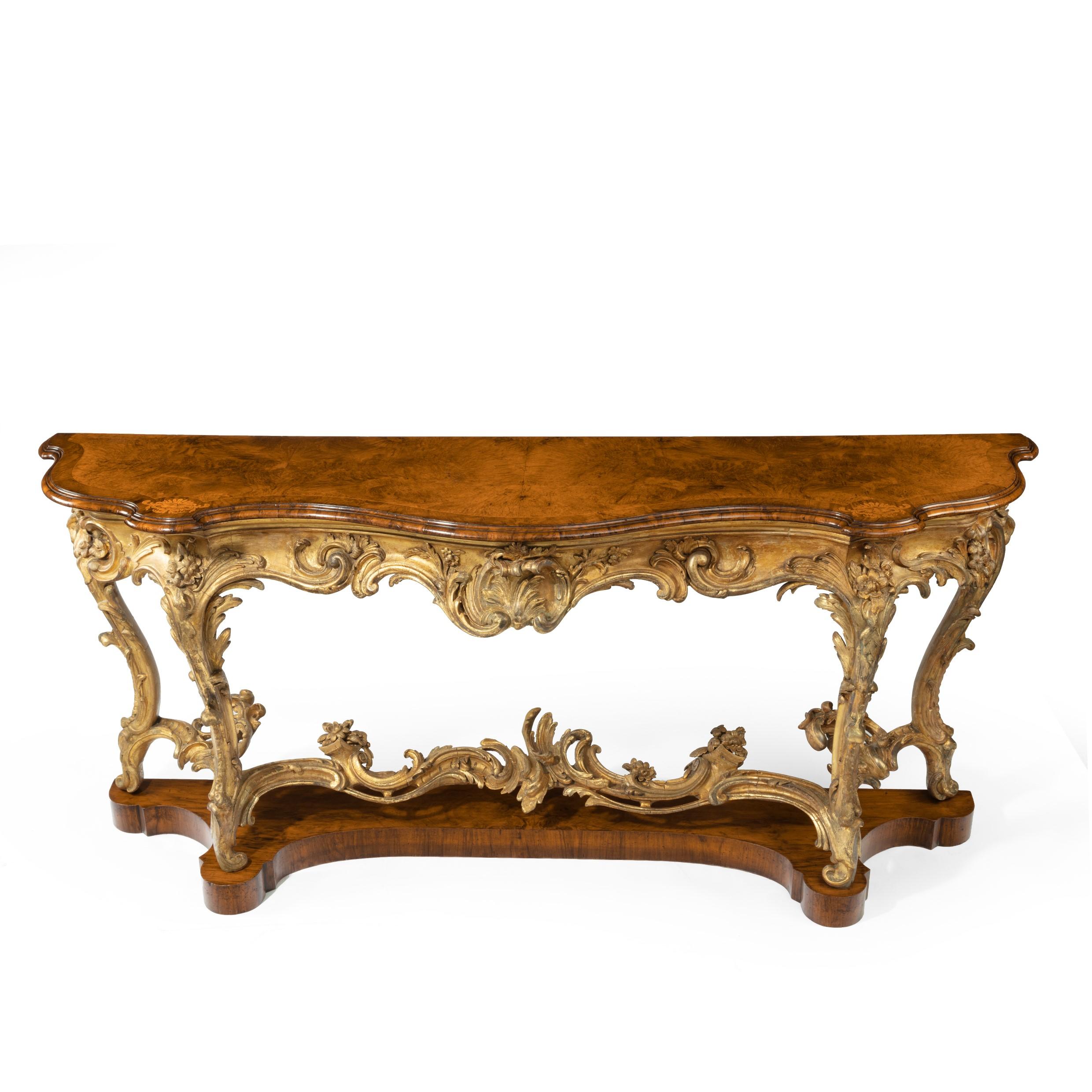 A mid Victorian wood and gilt console table, the shaped top with an amboyna field within wide walnut cross-banding inlaid with boxwood lion’s masks, raised upon a flamboyant gilt gesso base in the rococo taste with C-scrolls, acanthus leaves,