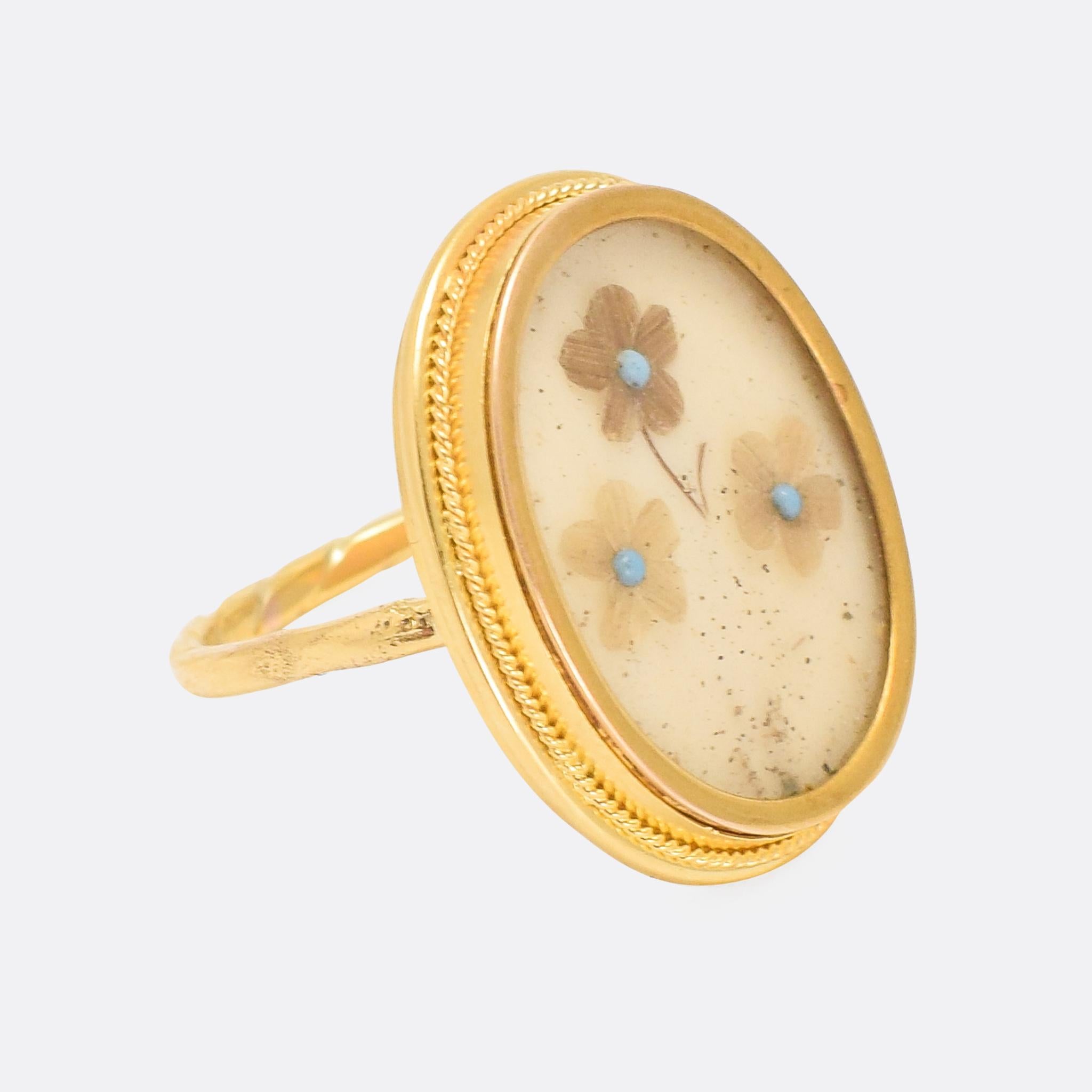 A weird and wonderful antique memorial ring dating from the mid Victorian era. Behind a glass panel are three forget-me-not flowers, with turquoise centres and hair-work petals. It's crafted in 18 karat gold, with a ropework trim around the head and