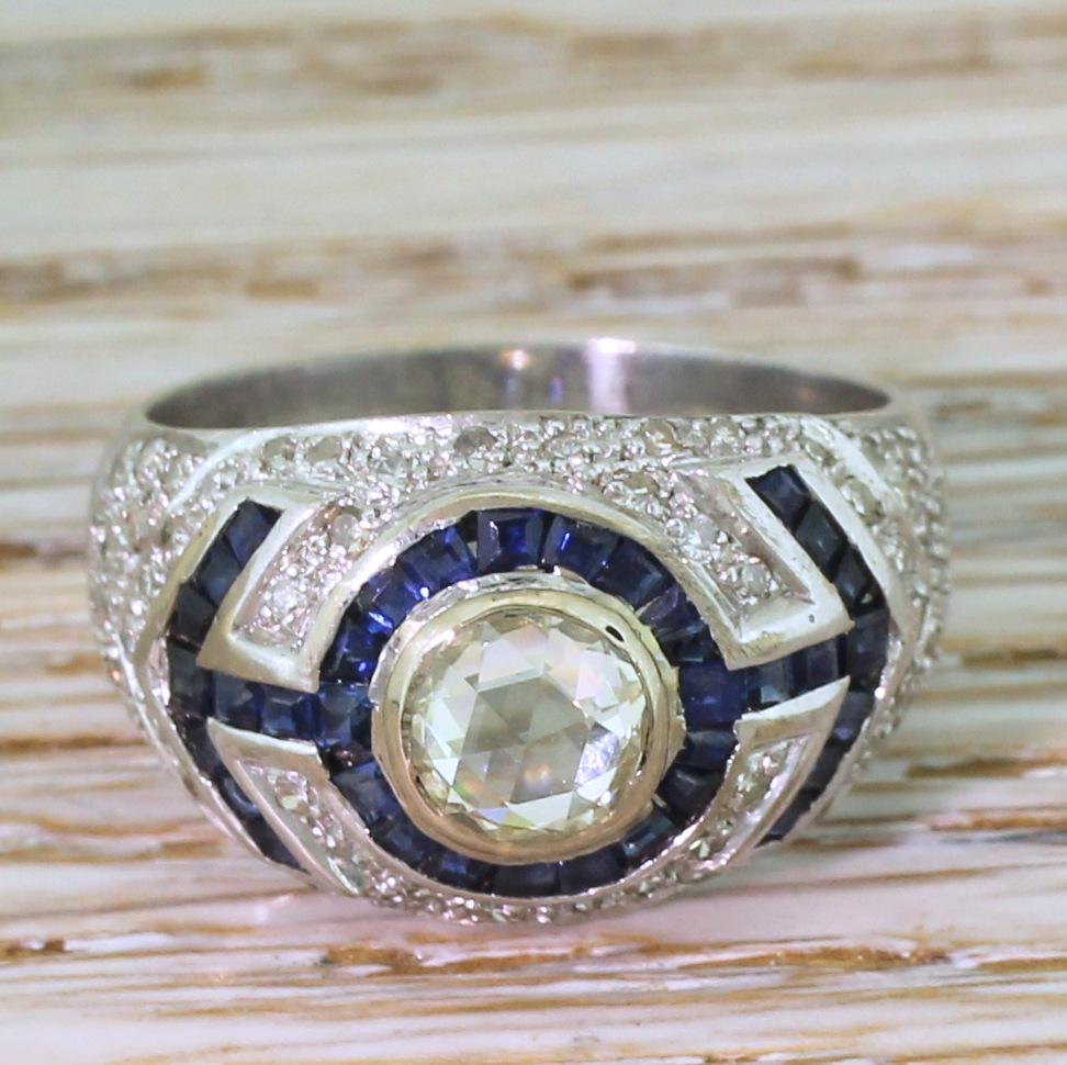 A wonderful rose cut diamond and sapphire ring. The impressive, high domed rose cut features twenty-four facets, creating a soft and diffused sparkle. The centre stone is rubover set and surrounded by a border of vibrantly blue step-cut sapphires,