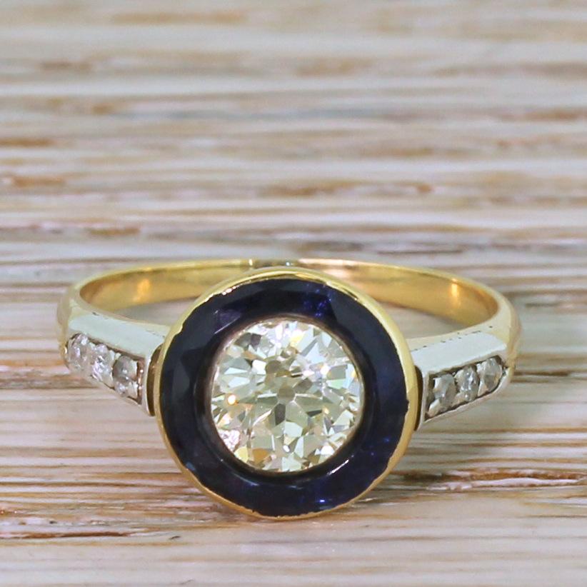 A super beautiful and undeniably unique ring. The old European cut diamond in the centre is internally clean and displays a very soft yellowish hue. Extraordinarily, the diamond is fully embedded within a round, deep blue sapphire which is itself