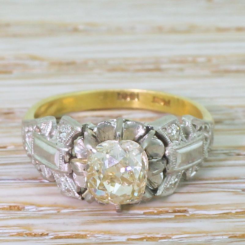 A classy and unique vintage engagement ring. The old mine cut diamond in the centre displays a distinct bright yellow, and is secured in an eight-claw open collet above a scalloped back-plate. The bold, dramatically shaped platinum shoulders feature