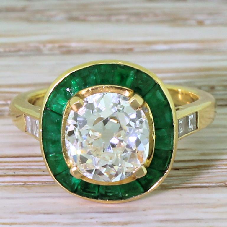 Incredible. This most beautiful of rings features a glowingly white and internally clean cushion shaped old cut diamond in the centre, encircled bright, verdant green calibré cut emeralds. Neat “V” detailing in the gallery leads to a yellow gold