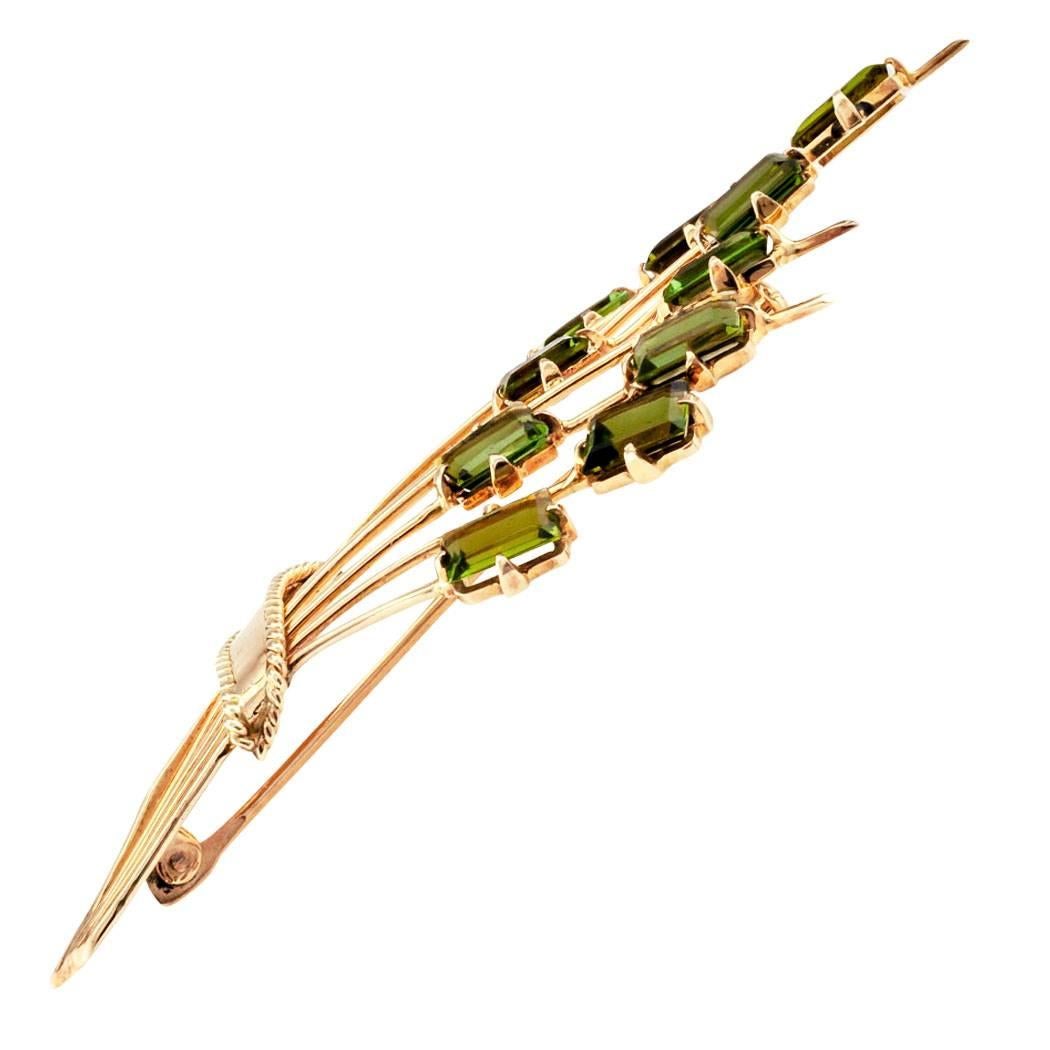 Mid century 1950s tourmaline and gold brooch. The hand fabricated jewel has been designed as a spray of ten rectangular-cut green tourmalines, each supported by curving stems, all bunched by an undulating ribbon of corded gold, mounted in 14-karat