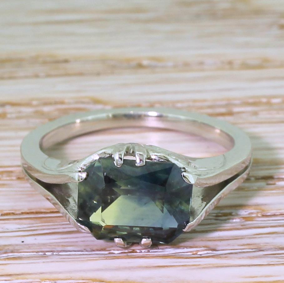 A fantasticly unique sapphire solitaire ring. The emerald cut sapphire in the centre is a strikingly unusual greenish blue, and is set laterally on the finger in gorgeously crafted white gold ring. Neat swirling detailing in the gallery leads to