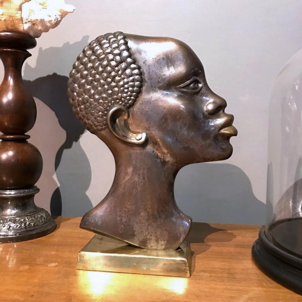 Schöner Bronzekopf einer afrikanischen Frau aus der Mitte des Jahrhunderts, hergestellt in Österreich in den 1950er Jahren, mit einem Ohrring aus Messing und einem Sockel aus Messing im Stil der Hagenauer Werkstatte. 
In sehr gutem Zustand mit