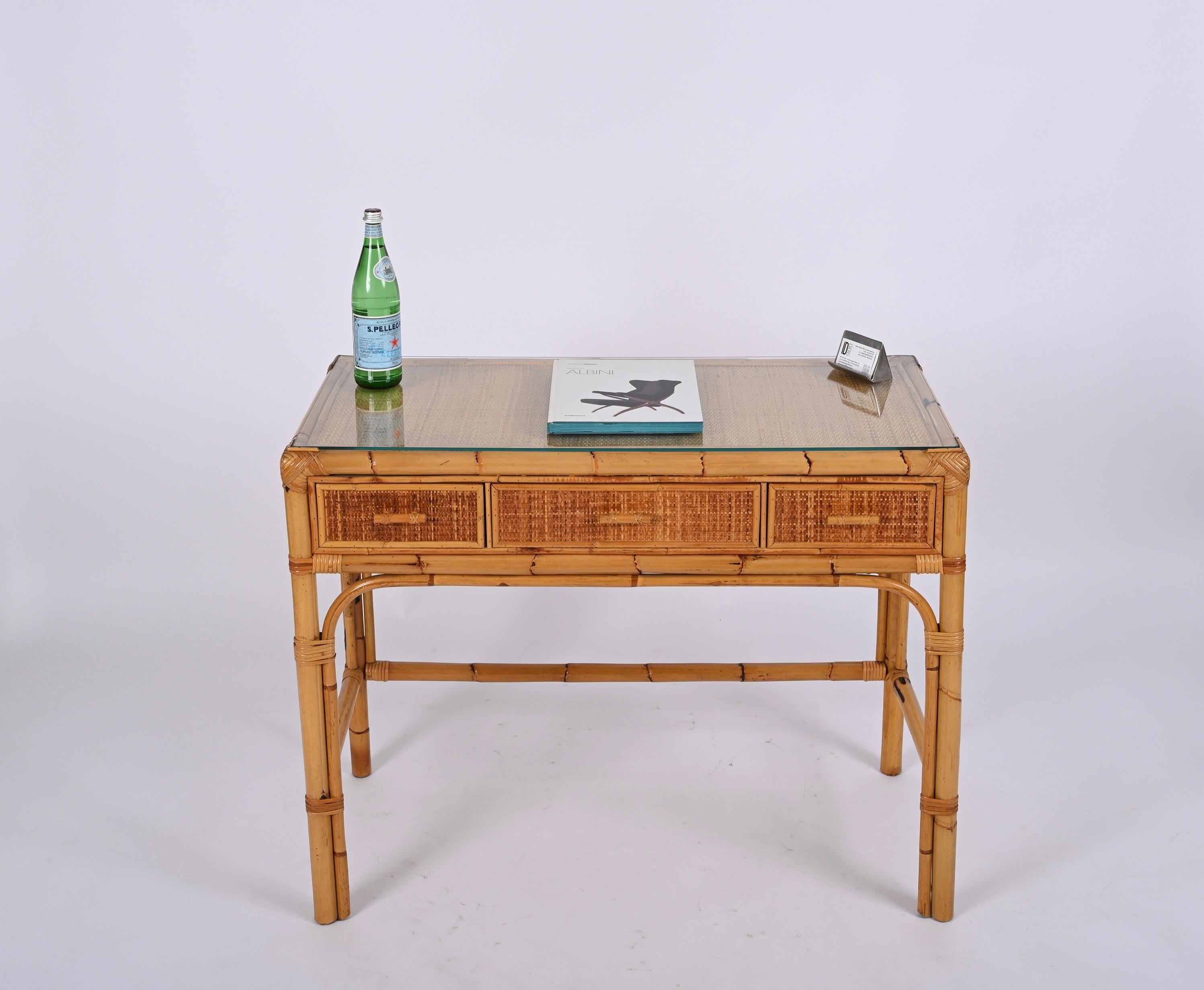 Unique midcentury rectangular bamboo and rattan wicker desk with crystal glass top and three drawers. This excellent piece was designed in Italy during the 1970s.

This stunning desk features a structure in curved bamboo cane enriched by woven