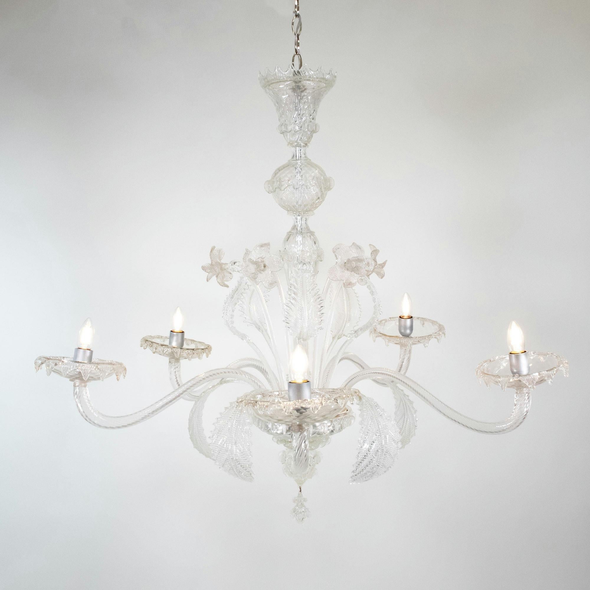 Questo lampadario artigianale in Cristallo di Murano ha cinque bracci a volute ondulate e presenta una colonna centrale a bulbo con dettagli naturali che spuntano, qui stilizzati con narcisi e foglie. I bracci terminano con coppe poco profonde
