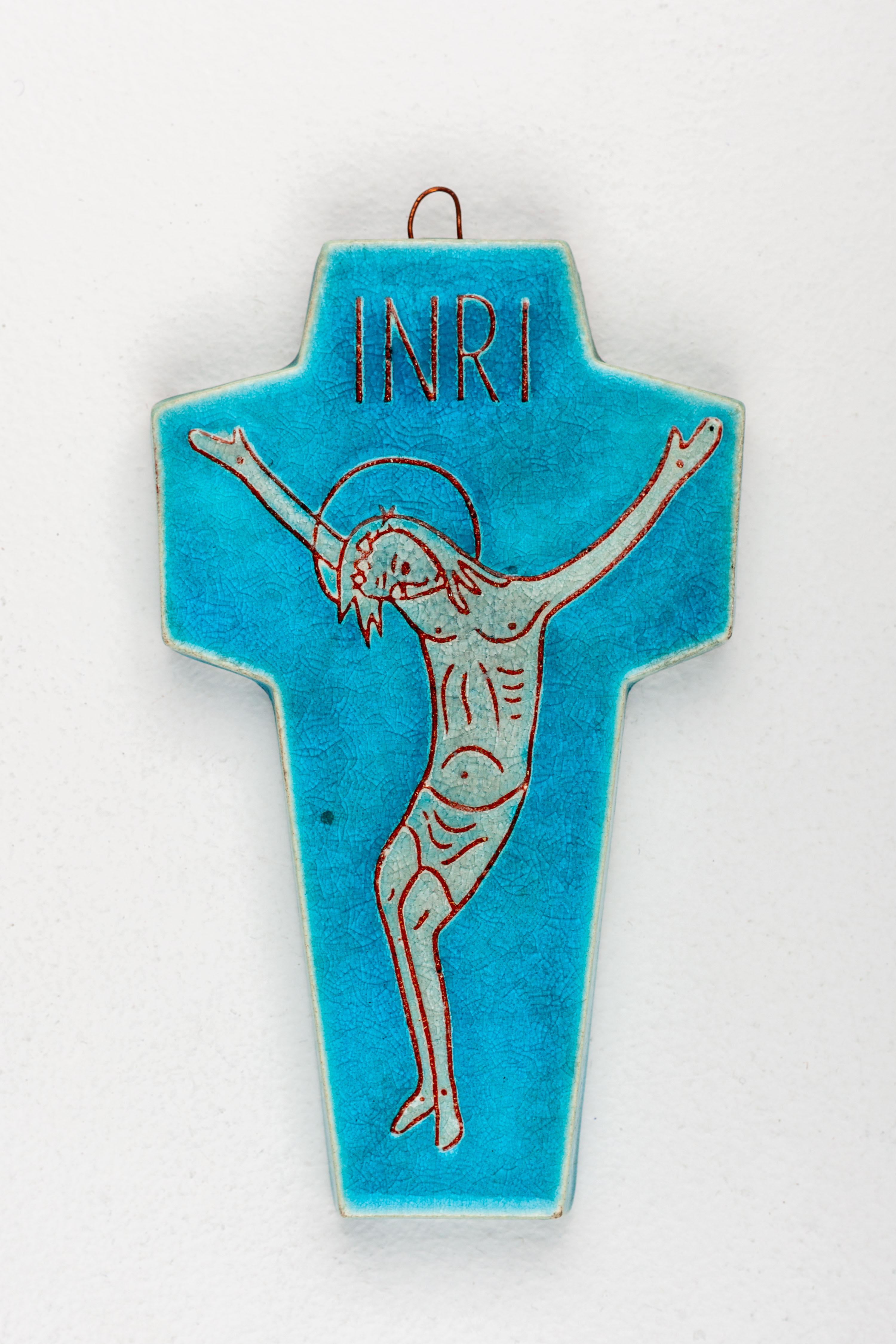 This blue-green ceramic wall cross, handcrafted in Europe by a Studio Pottery artist, blends modernist aesthetics and traditional iconography. The cross features a wide body with short wings, adorned with a modernist depiction of Jesus Christ. Above