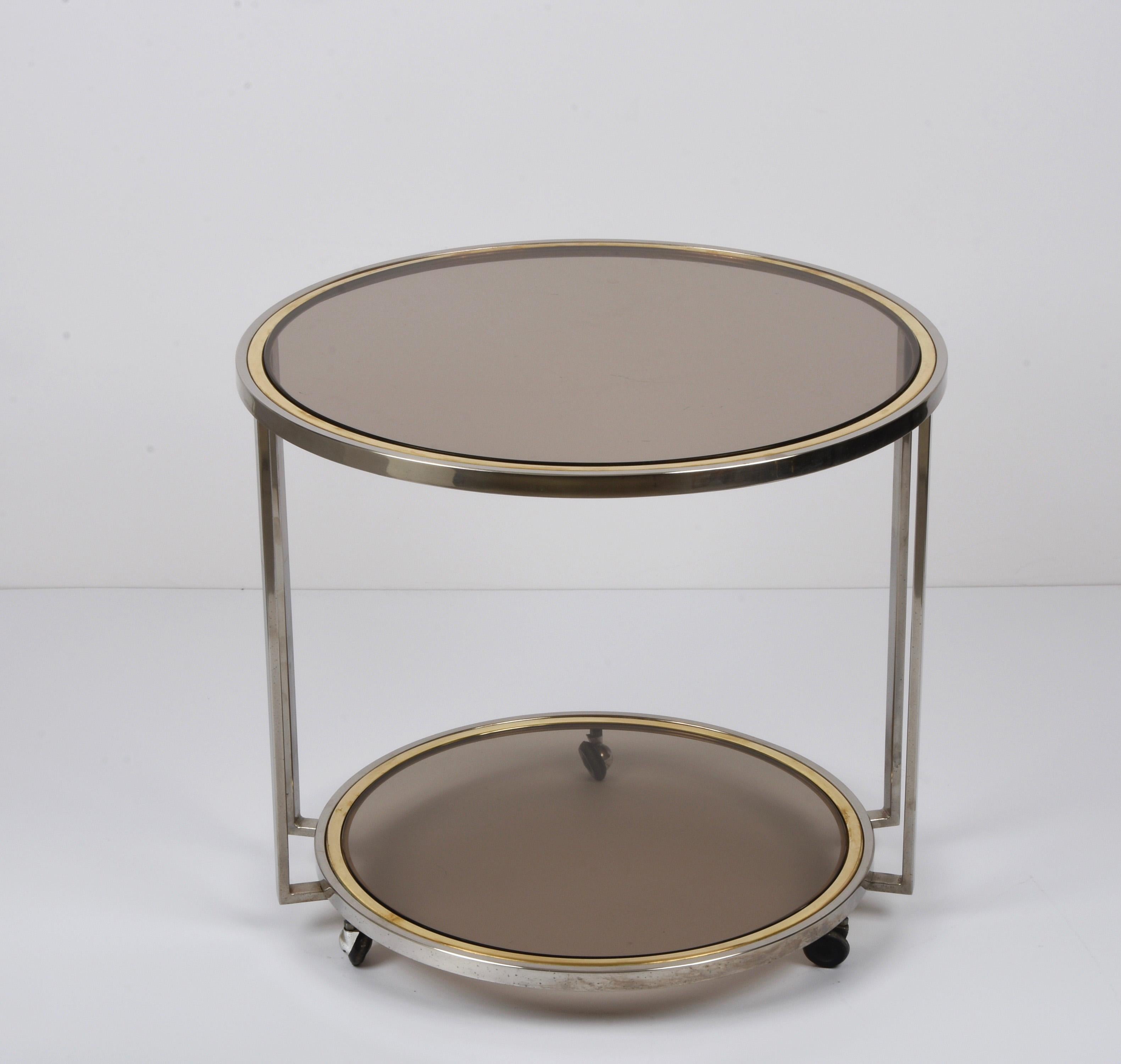 Incredible midcentury three-wheeled round side table in chrome, brass, and smoked bronzed glass. The upper floor has a diameter of 61 cm while the lower one is delightfully smaller and has a diameter of 51 cms. This wonderful piece was designed