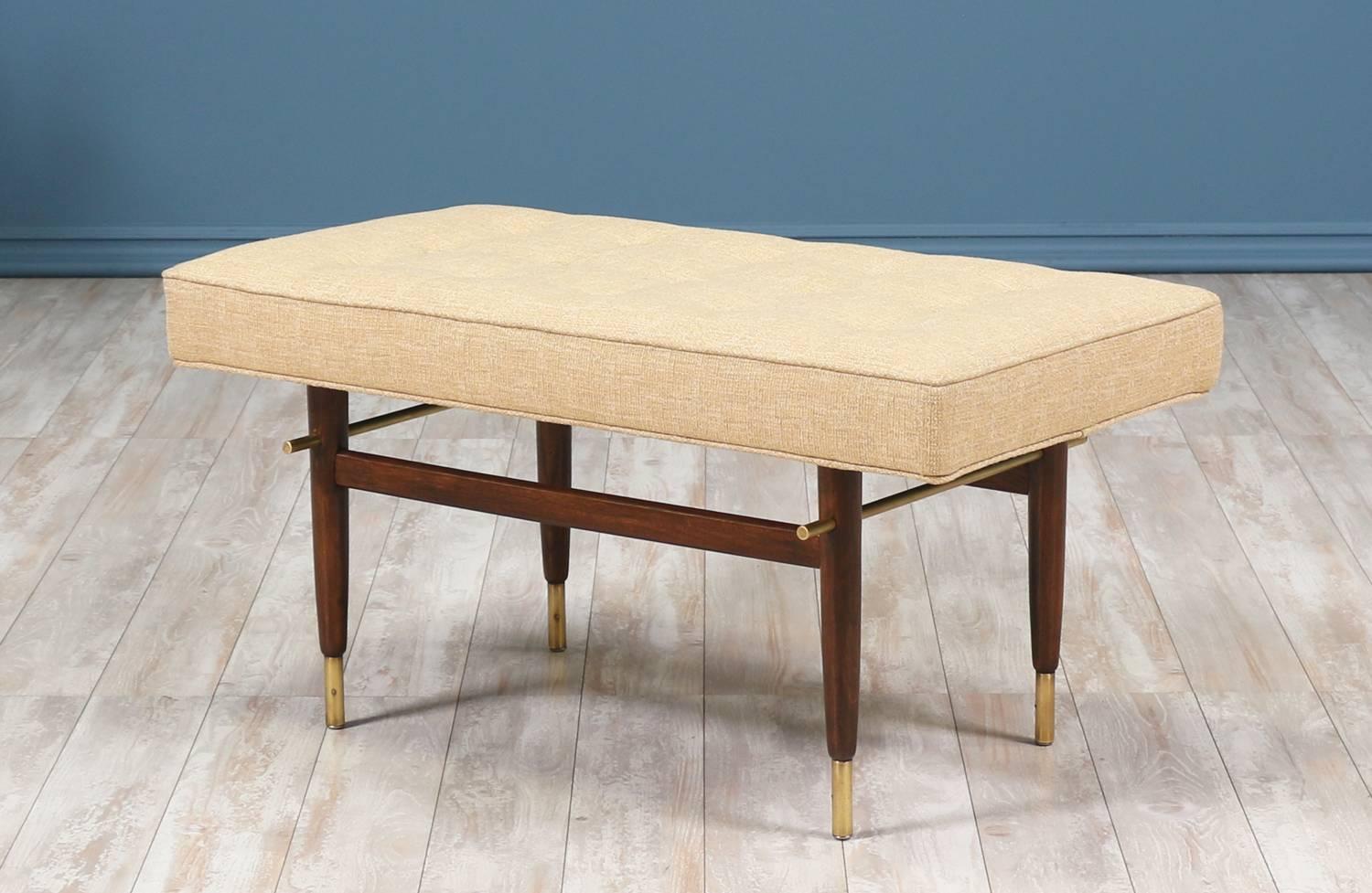 Mid-Century Modern tufted bench designed and manufactured in the United States, circa 1960s. This bench features a walnut wood base with two brass side stretchers and elegant brass sabots on all four tapered legs. The new high-density foam seat