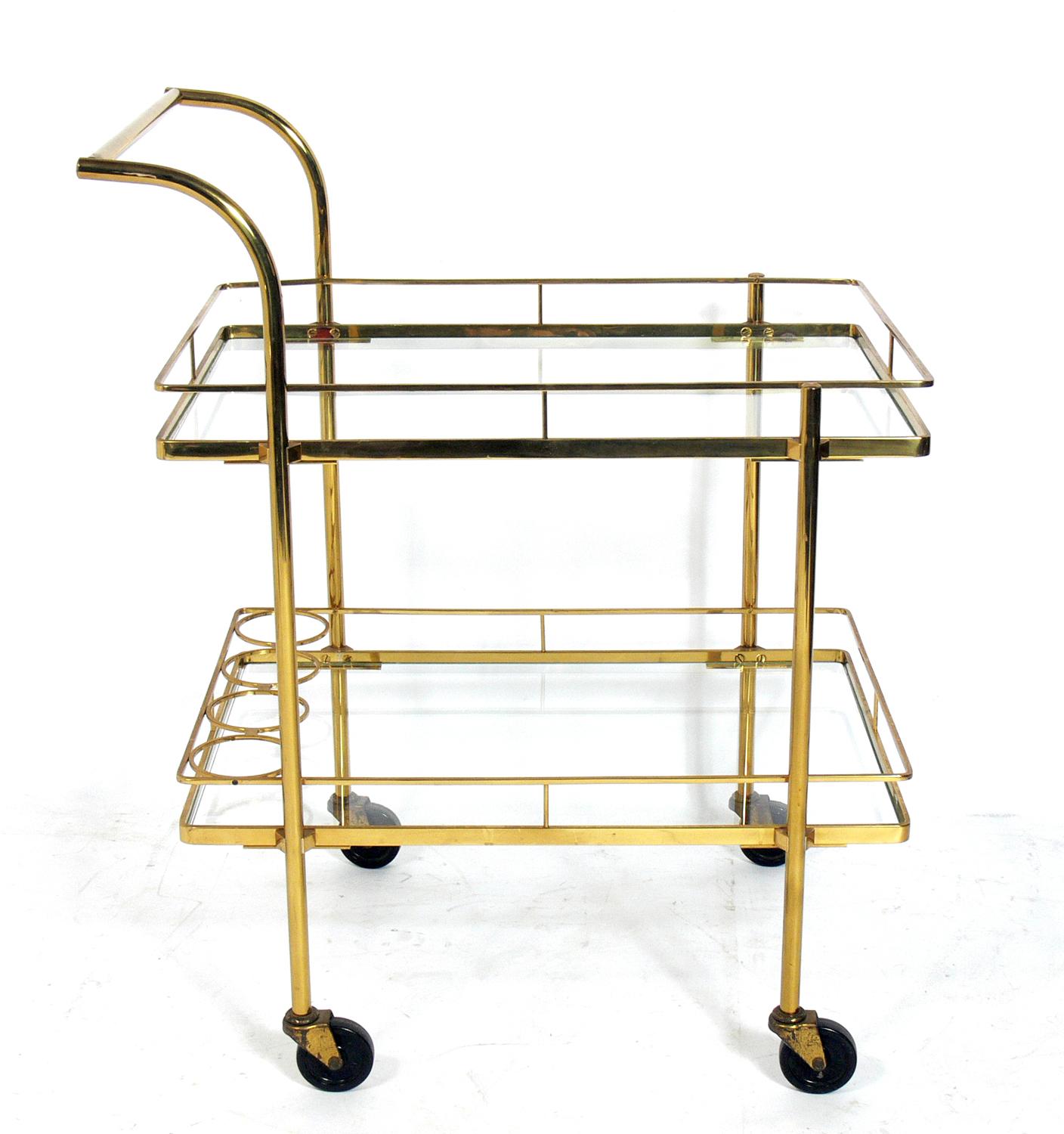 Midcentury brass bar cart, probably Italian, circa 1960s. It is a versatile size and can be used as a bar or serving cart or as extra rolling storage in an office. It has four bottle holders at the bottom and two large glass shelves.