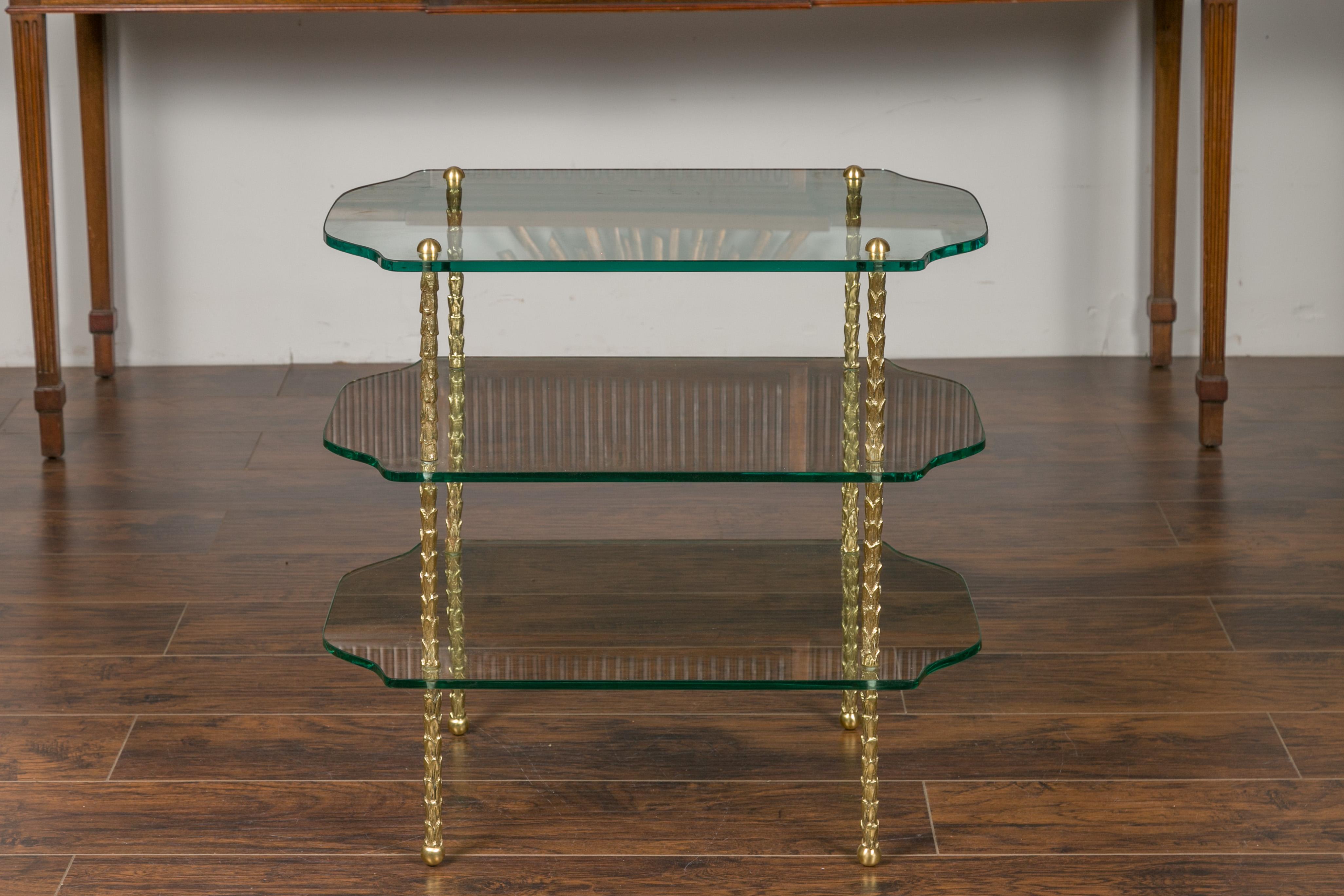 A vintage brass tiered table from the mid-20th century, with glass shelves and foliage motifs. Created during the midcentury period, this table features three shaped glass shelves secured by a brass structure adorned with stylized foliage motifs.