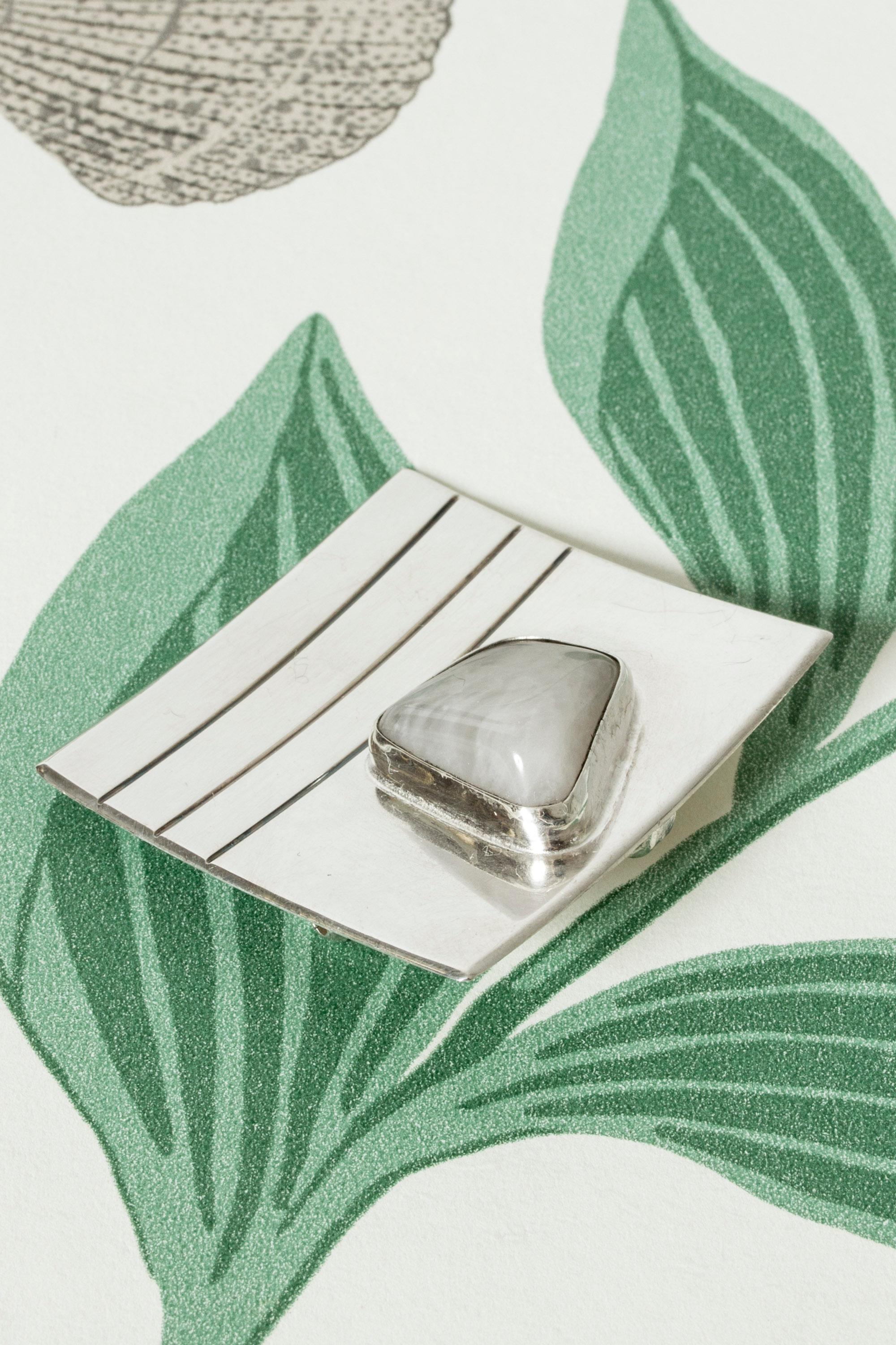 Cool, large brooch by Heikki Kaksonen, made from silver in a square form with embossed stripes. A smoothly tumbled moonstone makes a beautiful organic addition to the strict silver form.

