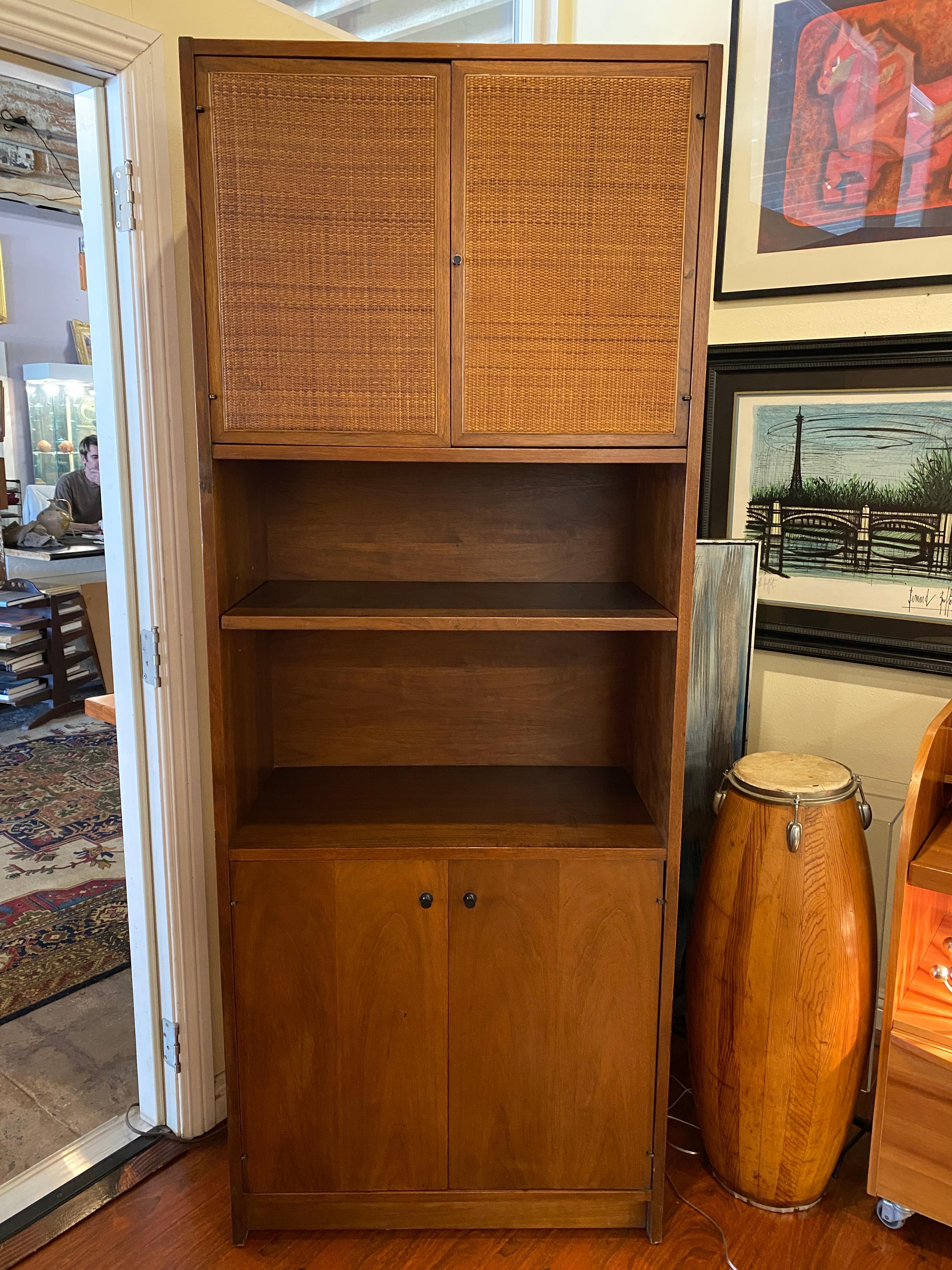 This tall cabinet, designed by Jack Cartwright for Founders Furniture around 1960, reflects the clean lines and functional elegance of midcentury modern design. Constructed from oiled walnut, it features upper doors with cane panels in excellent
