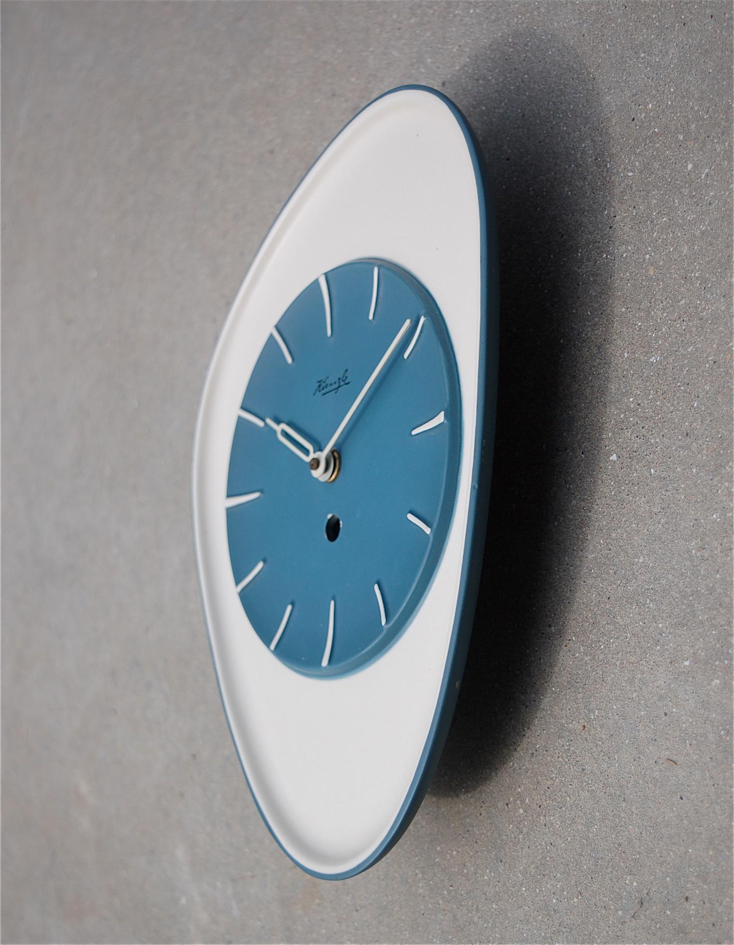 Mid-Century Modern ceramic wall clock by German quality manufacturer Kienzle. The design is quintessentially 1950s with its asymmetrical shape in a matte blue and white/cream glaze. The makers name is featured at the top of the dial. The hours are