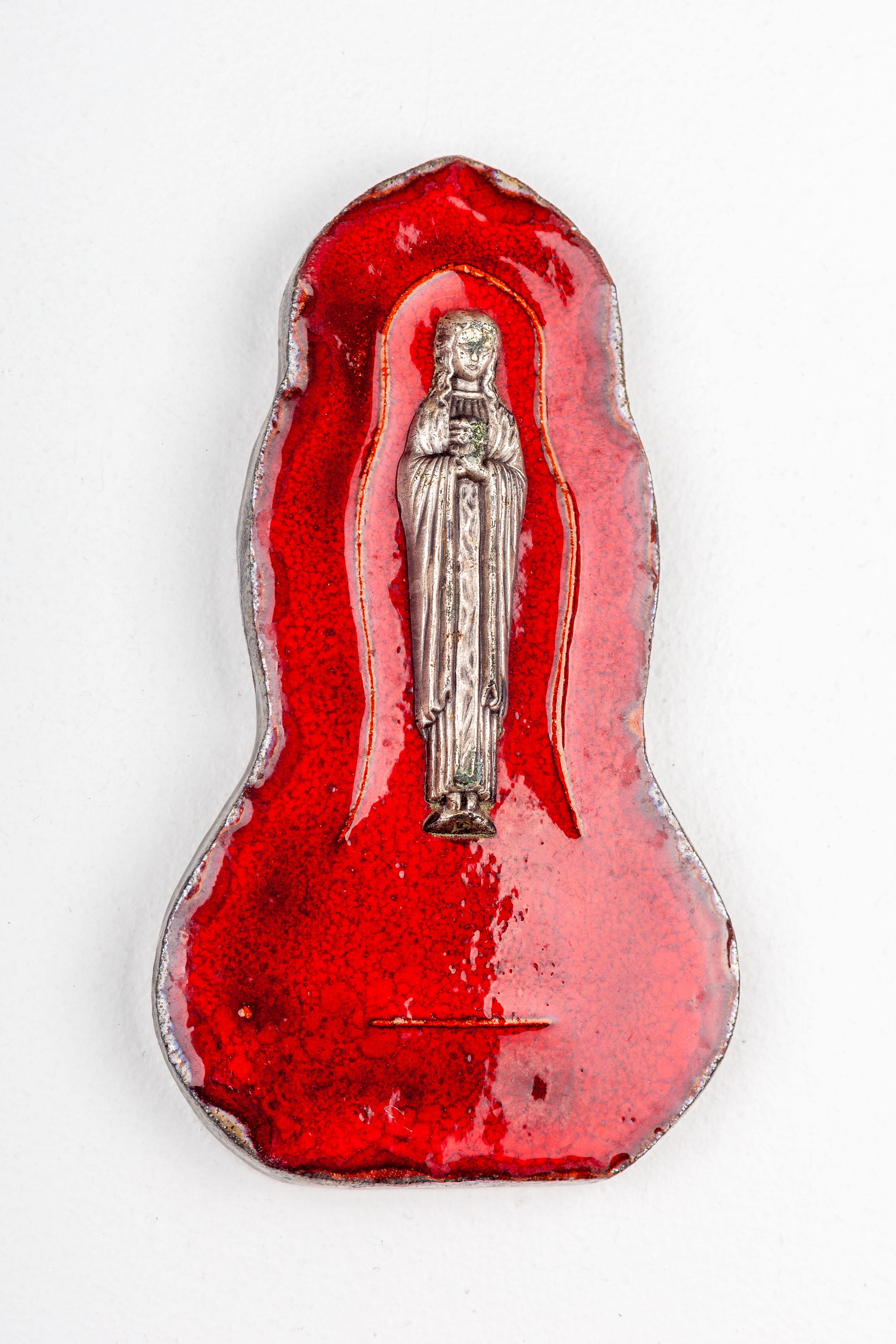 This midcentury ceramic wall plaque features a metal Virgin Mary statuette. The ceramic plaque is adorned with a painted red lava texture, evoking a sense of mysticism that mirrors the mental landscape of the Virgin Mary. The juxtaposition of the