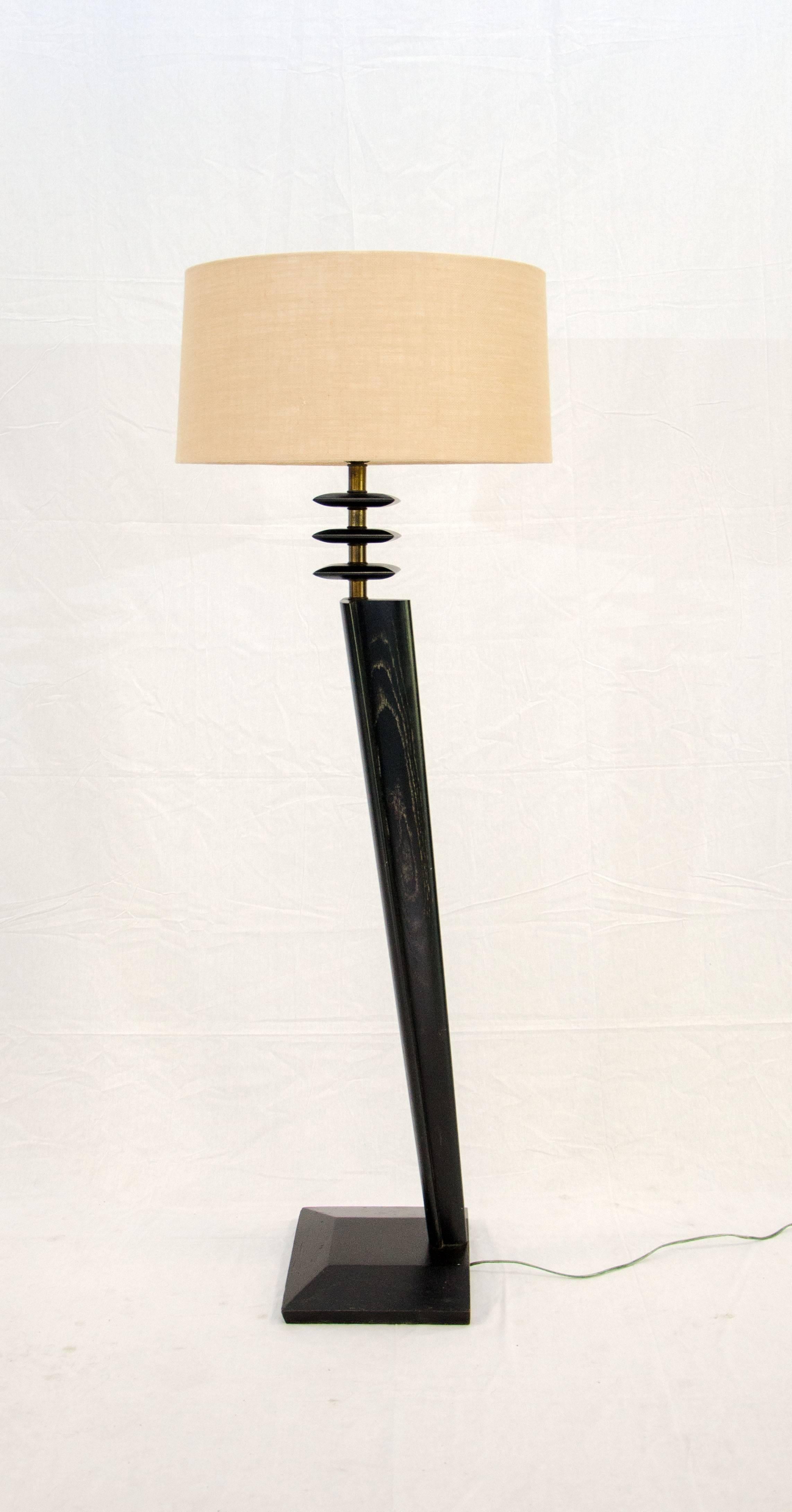 This floor lamp has an unusual angular design the main post slants upward from just one side of the rather than being centered on the base. The lamp is accented by three square wooden pieces with a brass post between them. The new lightweight burlap