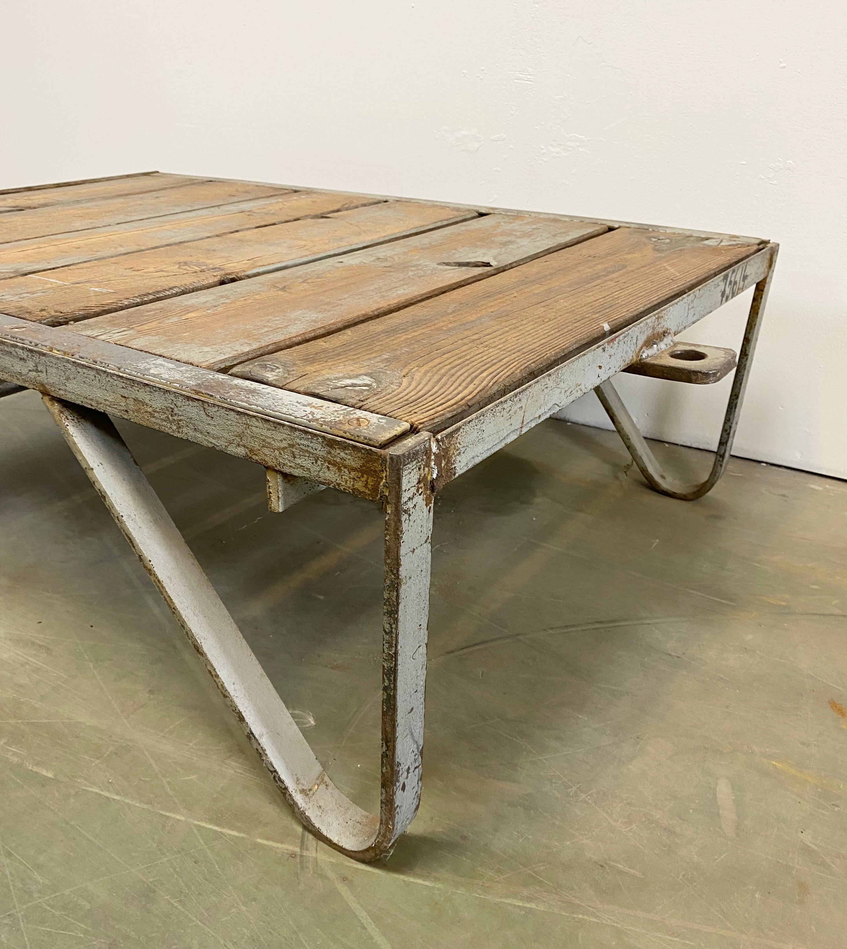 Former pallet truck from a factory now serves as a coffee table. It features an iron construction with two wheels and a solid wooden plate. The weight of the table is 35 kg.