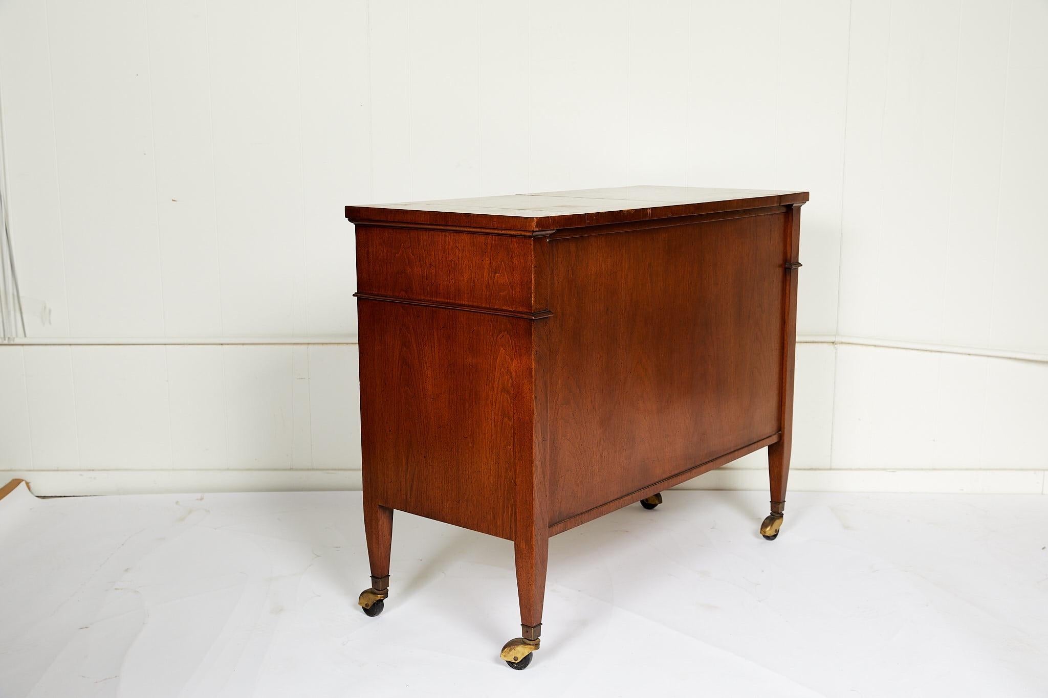 This 20th century vintage sideboard features a walnut finish on all sides, convertible black formica top, and casters for versatility and easy service. The case holds one wide drawer with brass ring pulls above two Regency style paneled doors with