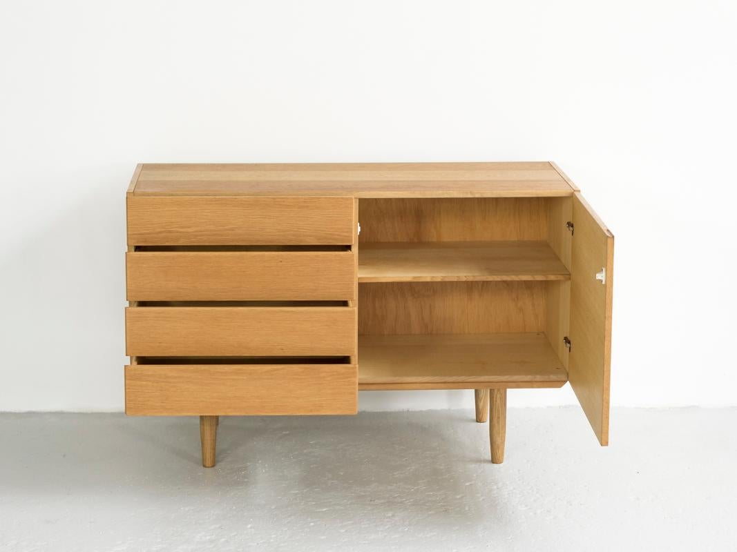 This midcentury cabinet is designed by Ib Kofod-Larsen and made in Denmark in the 1960s. It is a practical cupboard with 4 drawers and 1 door. The cabinet has integrated door and drawer handles. It is made of oak. There is a natural color difference