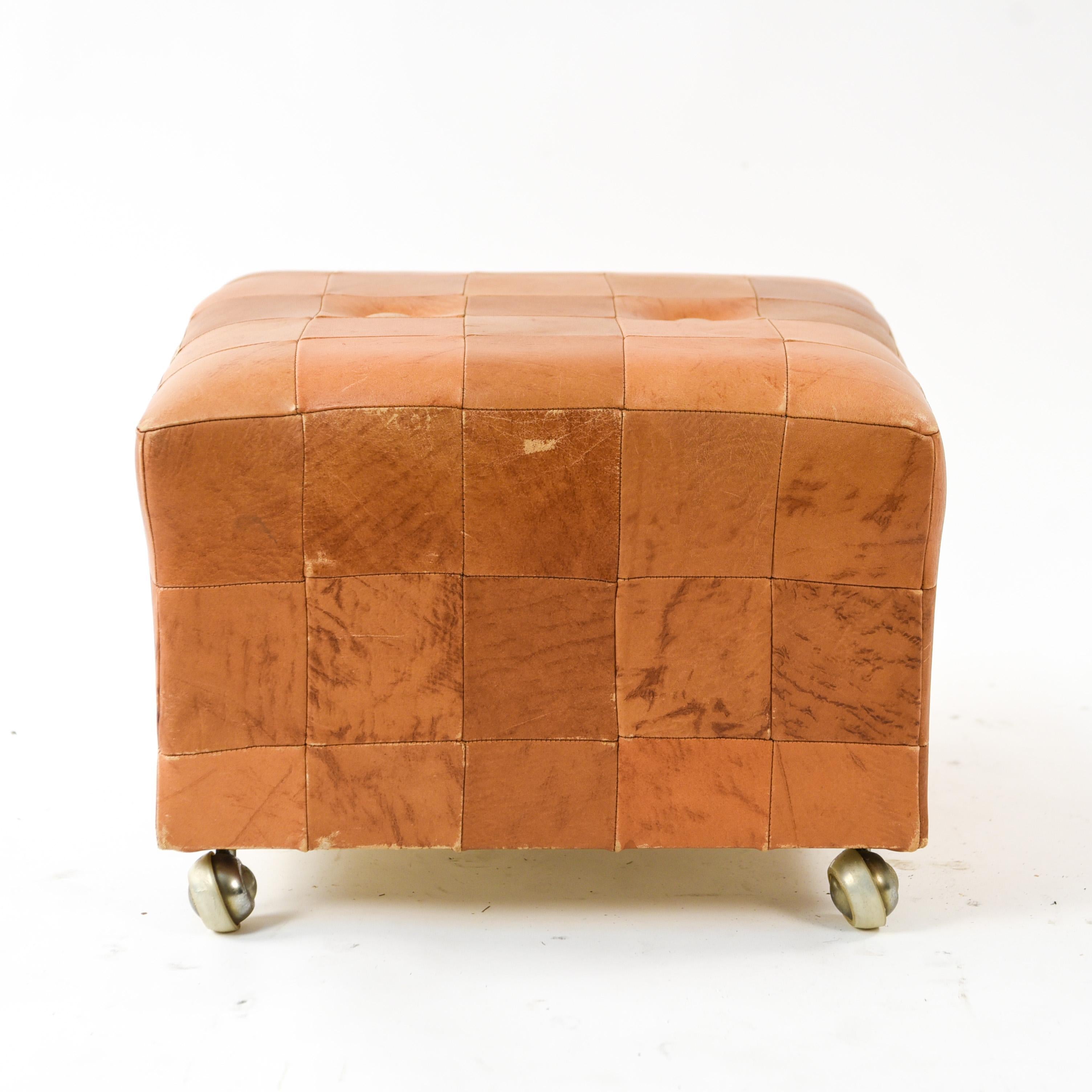 This midcentury cube ottoman is upholstered in butterscotch leather and features two button tufts on top. The upholstery creates squares over the surface of the ottoman which provides an aspect of visual interest. This piece is on casters which