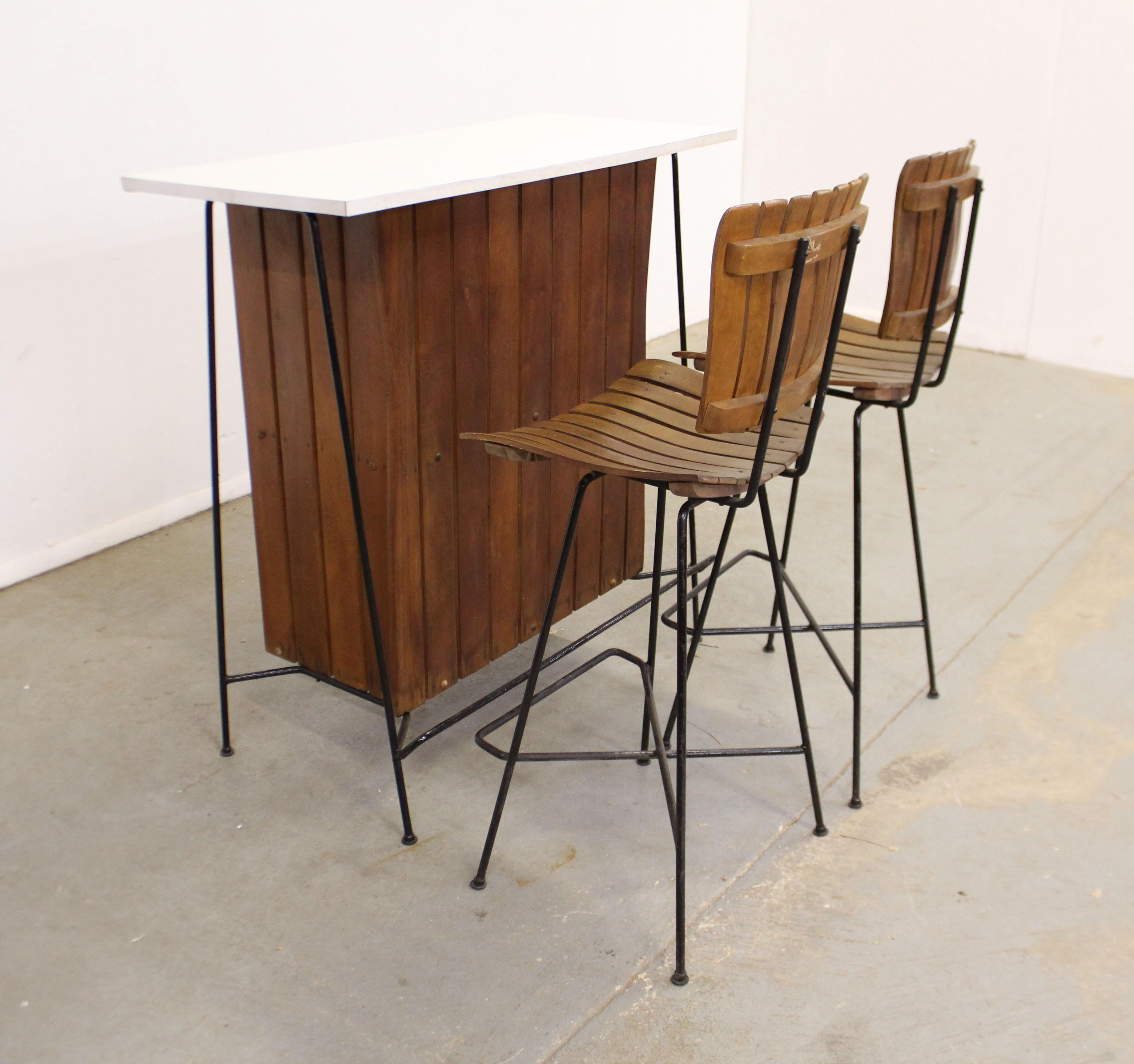 Offered is a 3-piece slat wood dry bar set designed by Arthur Umanoff. Includes two slat stools and a dry bar. The bar has a Formica top with rear shelving and wrought iron legs and foot rests. The set is in very good condition for its age, showing