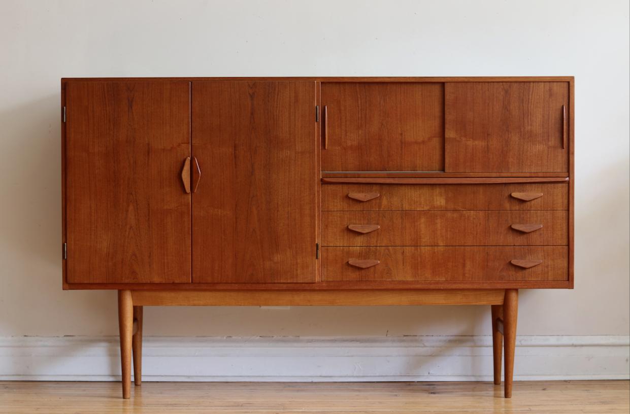 Mid-Century Modern Danish tall teakwood credenza.
Just imported from Copenhagen!
Beautiful arrow shaped handles and lovely teakwood grain.
Left features large cabinet space with adjustable shelving.
Right features a mirrored bar cabinet with