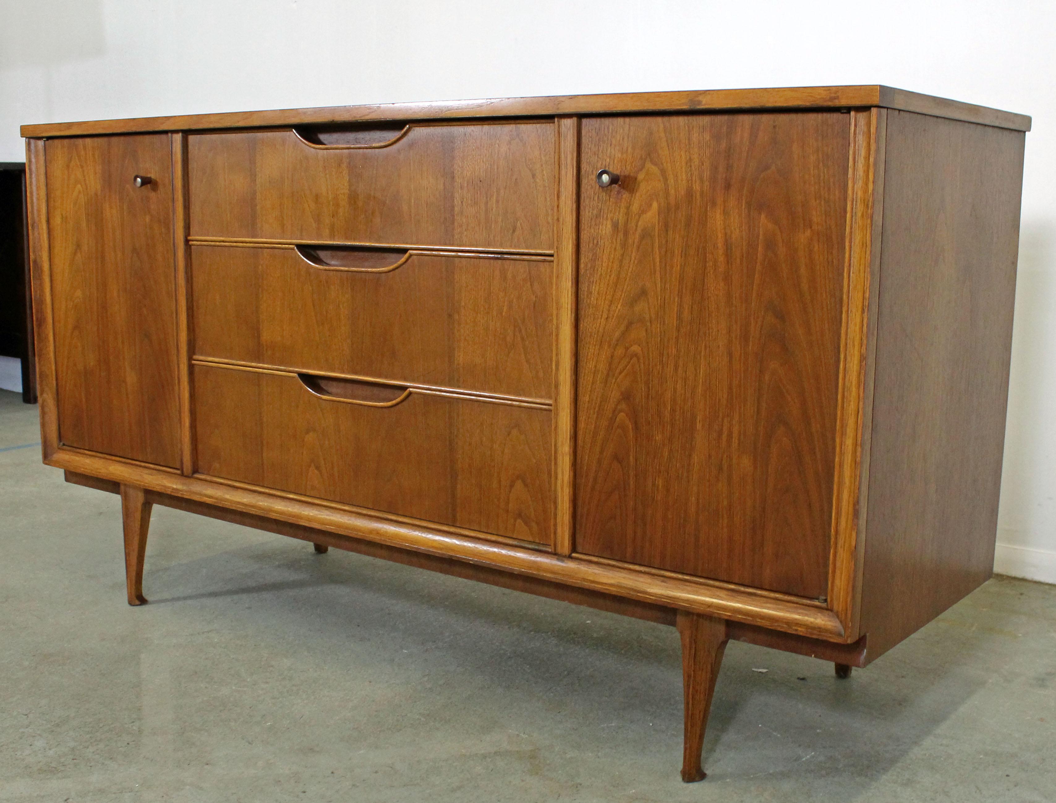 Offered is a Mid-Century Modern walnut credenza. Features two doors on both sides with storage/shelving inside, three centre drawers (top drawer has dividers), and recessed sculpted pulls. It is in good condition, shows some normal age wear (see