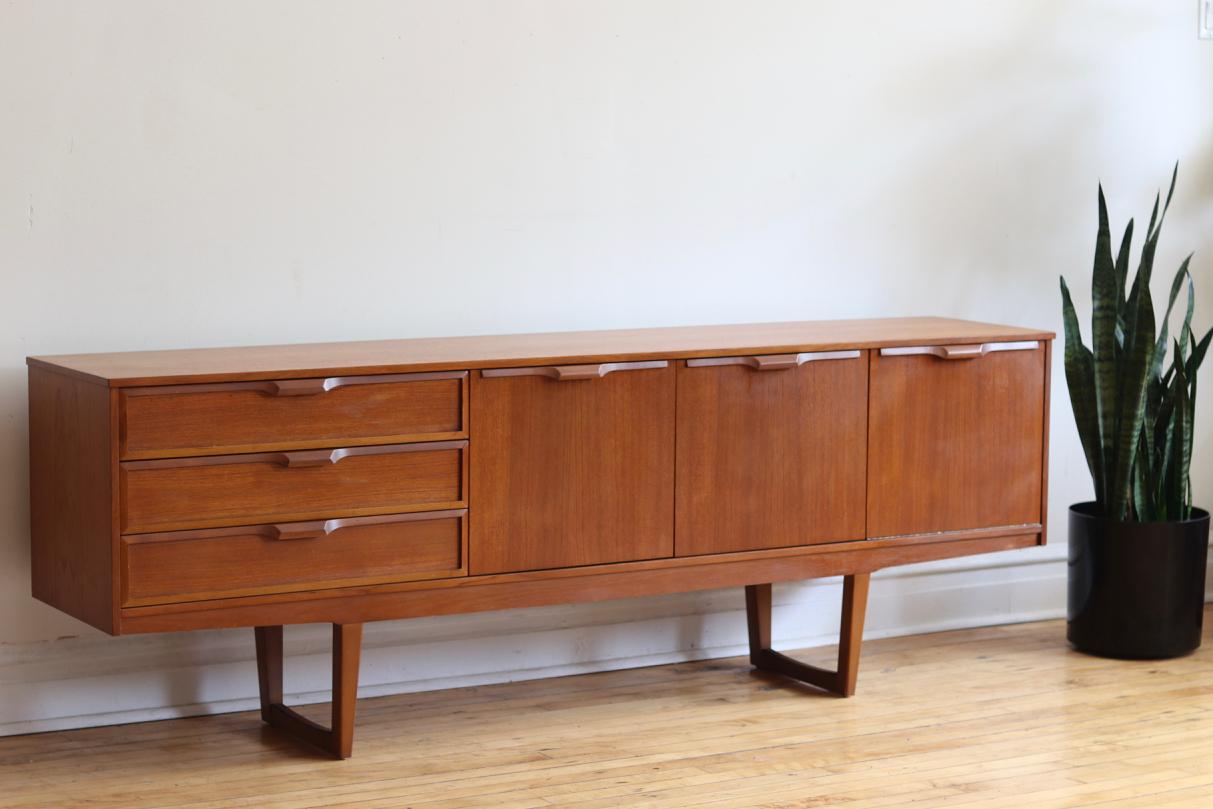 Mid-Century Danish modern teak wood sideboard featuring sled legs.
Made in England by S.F. Ltd.
Double cabinets in the centre.
Drop down bar cabinet on the right.
Three drawers on the left.
Removable shelving.
Beautifully