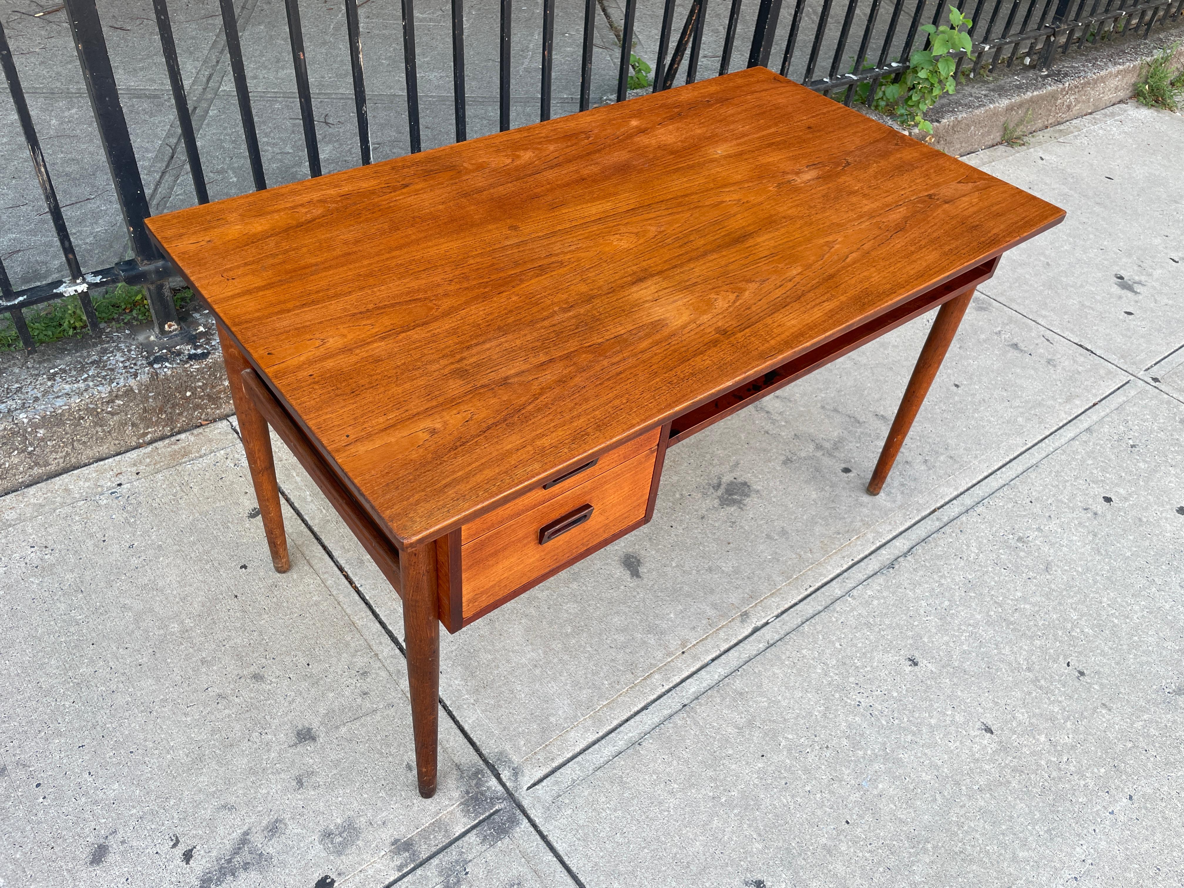 Great teak mid century Danish modern desk with 2 drawers with carved wooden handles. Danish design. Has a small shelf on the front side of desk and has a long open shelf that runs under the entire desk. made in Denmark. Located in Brooklyn NYC.