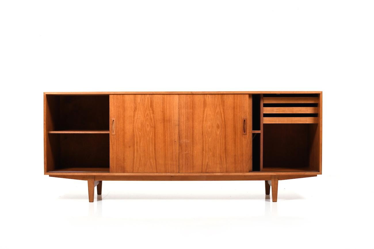 Midcentury Danish teak wooden sideboard. Front with four sliding doors. Behind the doors with shelves drawers. Standing on conical legs. Early 1960s. Beautiful teak color and veneer grain.