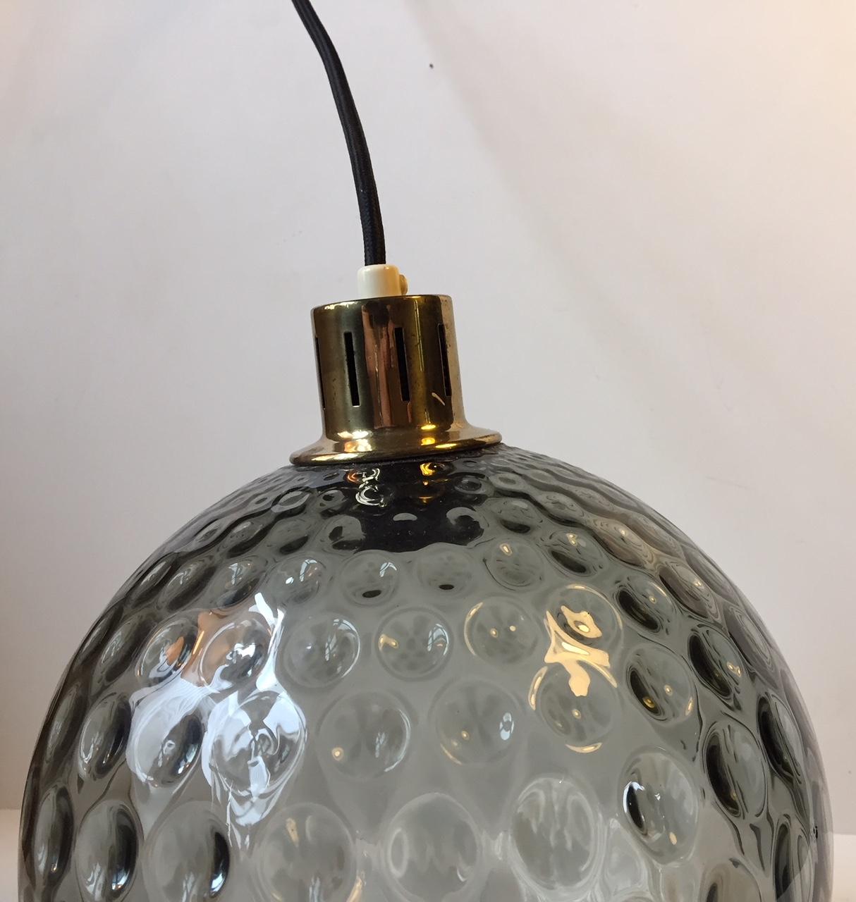 This pendant was manufactured by Orrefors in Sweden during the 1960s. It features an optical smoked glass dome and a partially perforated brass top. The design of this pendant is reminiscent of Wilhelm Wagenfeld and Venini/Murano.