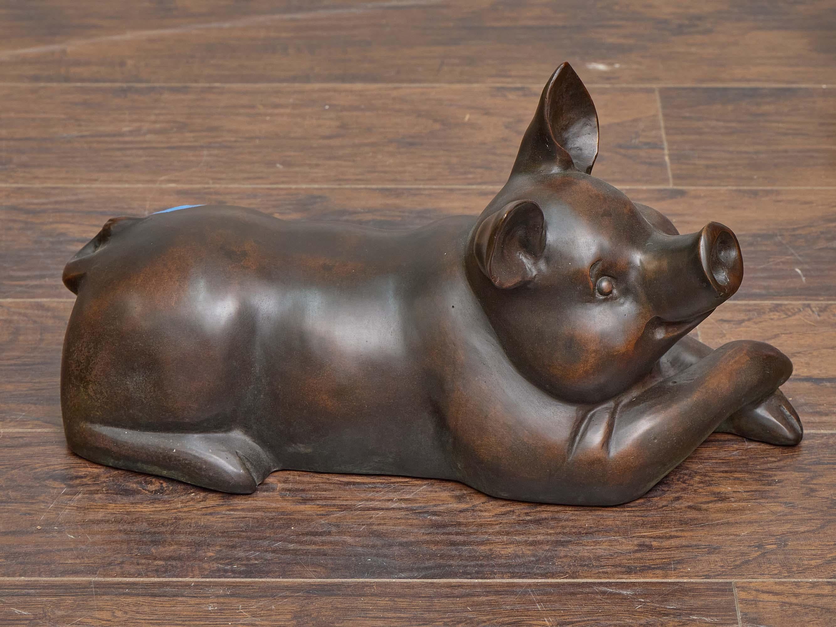 A delightful mid-20th century English bronze sculpture depicting a reclining pig, rendered with a balance of naturalistic detail and stylized charm. The pig is shown lying comfortably on its belly, with a slightly raised head and gently folded legs,