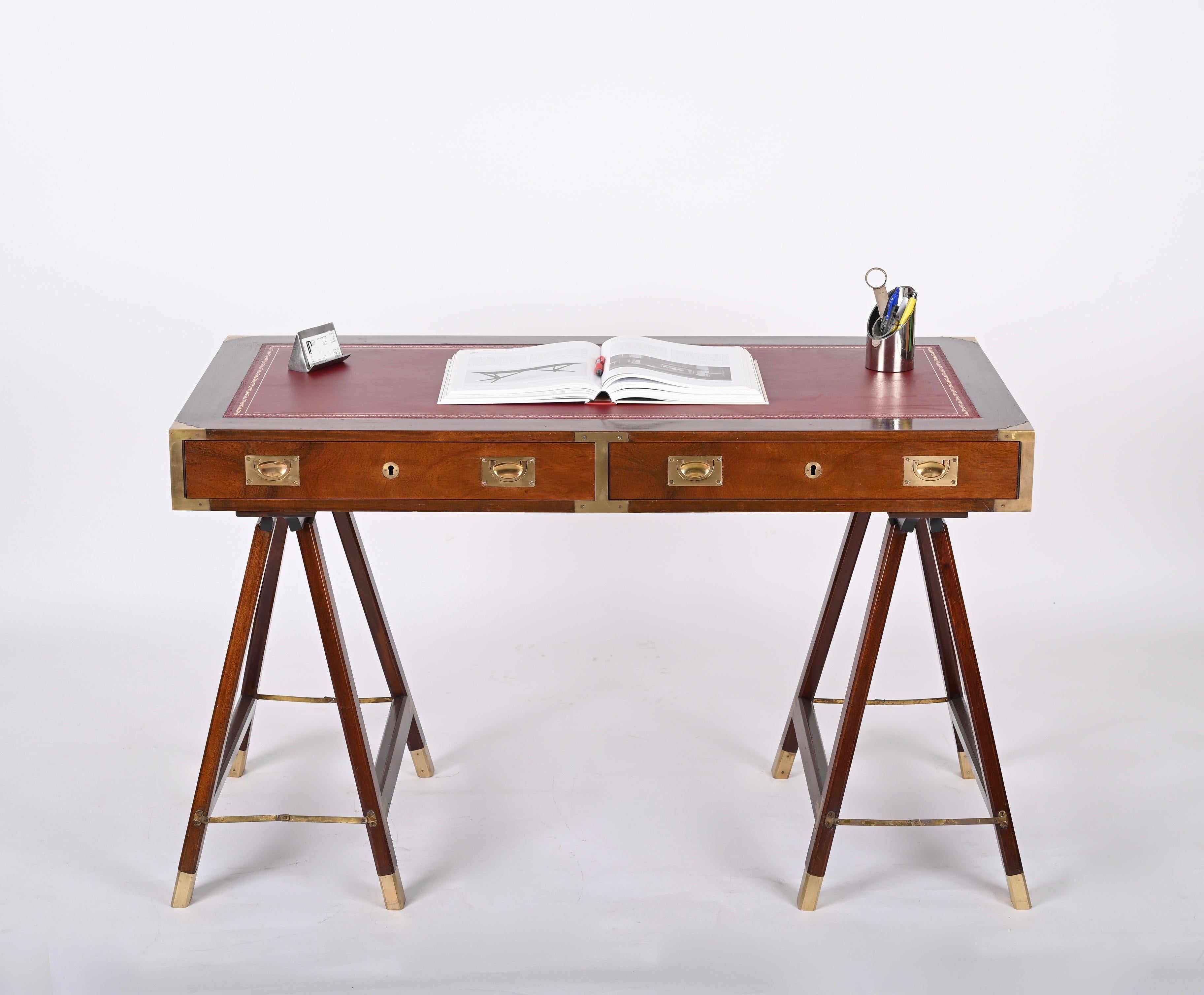Stunning midcentury desk in Victorian military style. This piece was produced in England during the 1950s-1960s. 
The desk is made in  walnut wood, the top is decorated with a beautiful red leather enriched by gilded ornaments. and brass finishes.