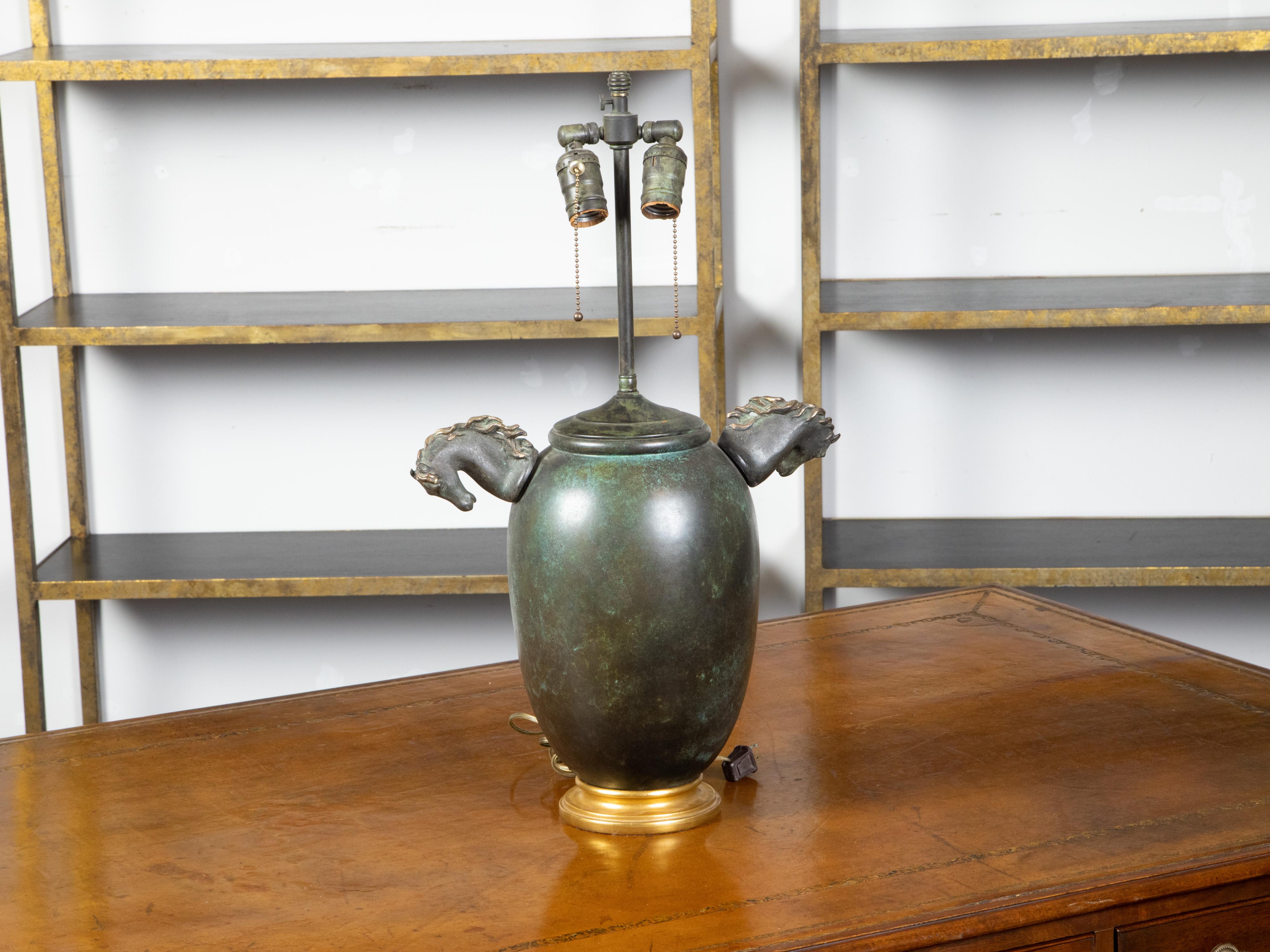 A vintage English bronze two-light table lamp from the mid 20th century with horse heads, gilded base and verdigris patina. Created in England during the Midcentury period, this table lamp features a generous bronze body boasting a verdigris patina,